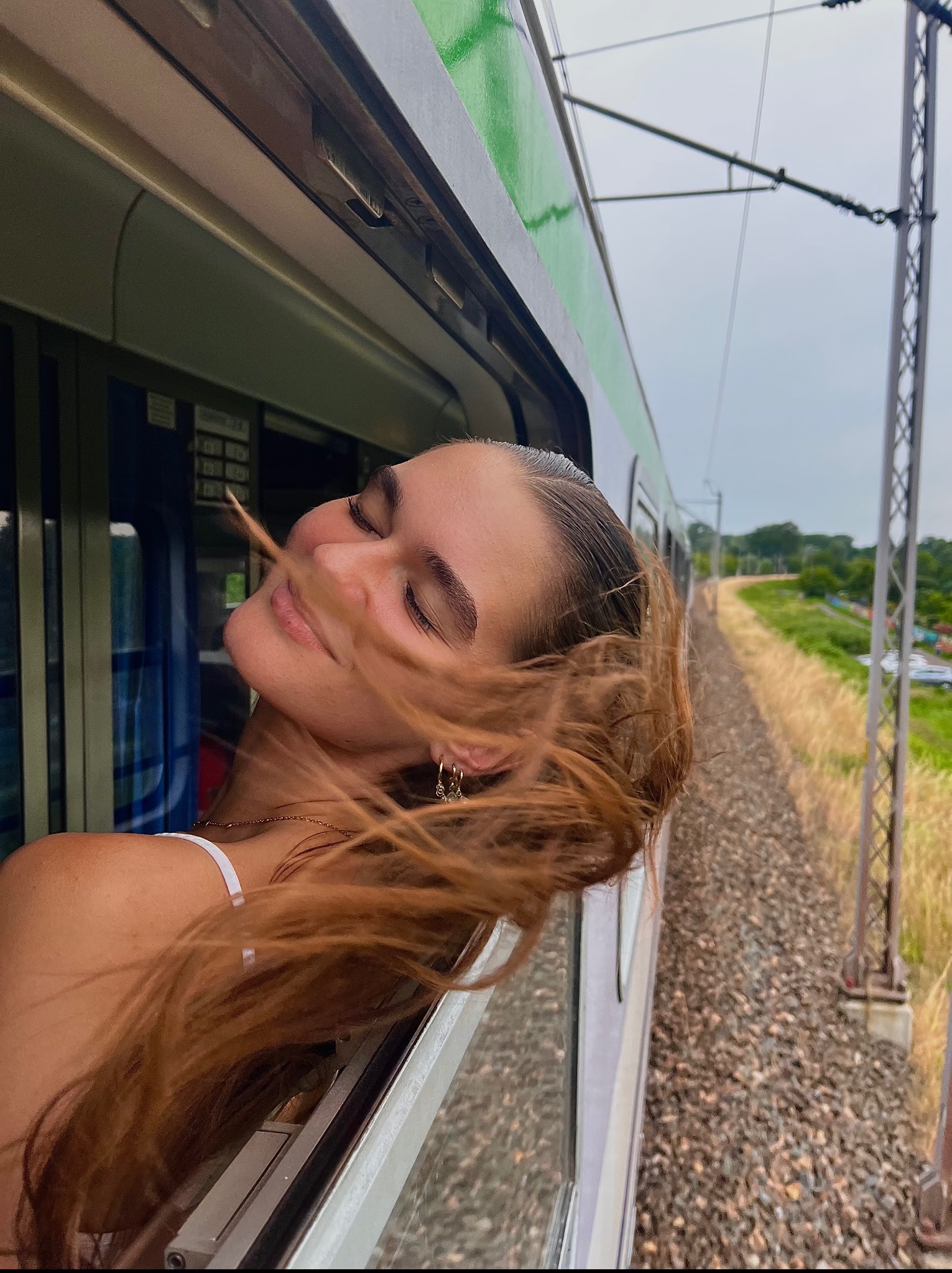 Uśmiechnięta, młoda kobieta z rozwianymi włosami wystawia głowę przez okno zielonego pociągu, w tle widoczne tory i krajobraz.