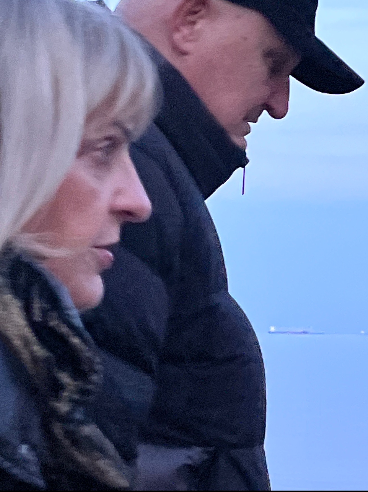 Profil kobiety z blond włosami i mężczyzny w czapce na tle błękitnego nieba i morza z widocznym statkiem w oddali. Ujęcie z bliska, skupione na twarzach.