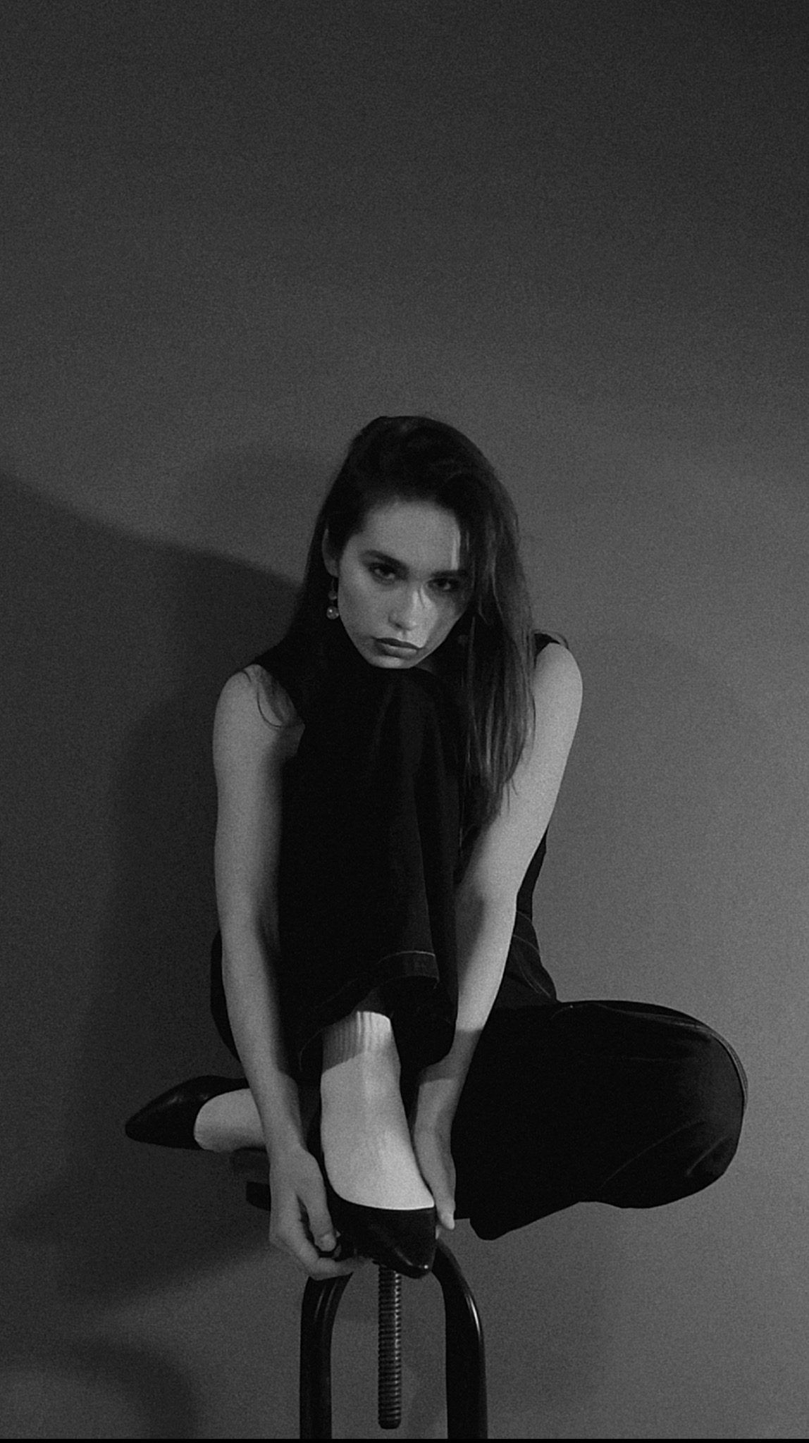 Monochromatyczne, studyjne ujęcie kobiety siedzącej na stołku, z nogami podciągniętymi do góry i dłońmi dotykającymi buta. Stonowana kolorystyka, minimalistyczne tło.