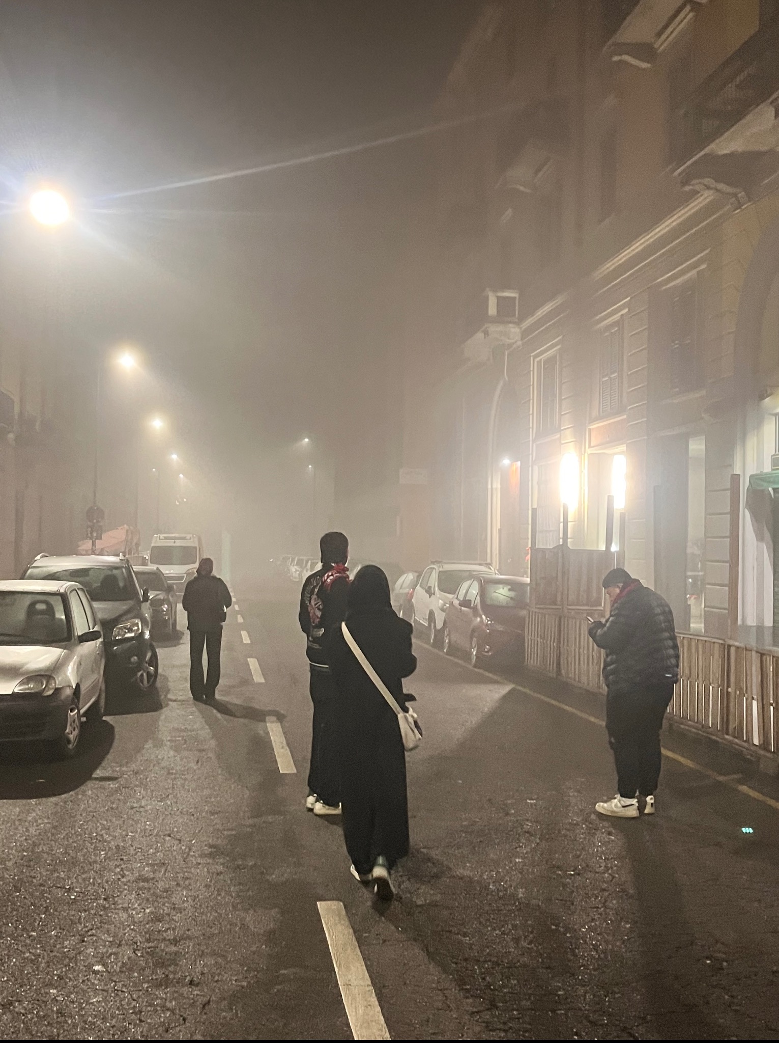 Nocna ulica w gęstej mgle z zaparkowanymi samochodami i sylwetkami przechodniów pod latarniami. Budynki po obu stronach drogi. Ujęcie z perspektywy pieszego.