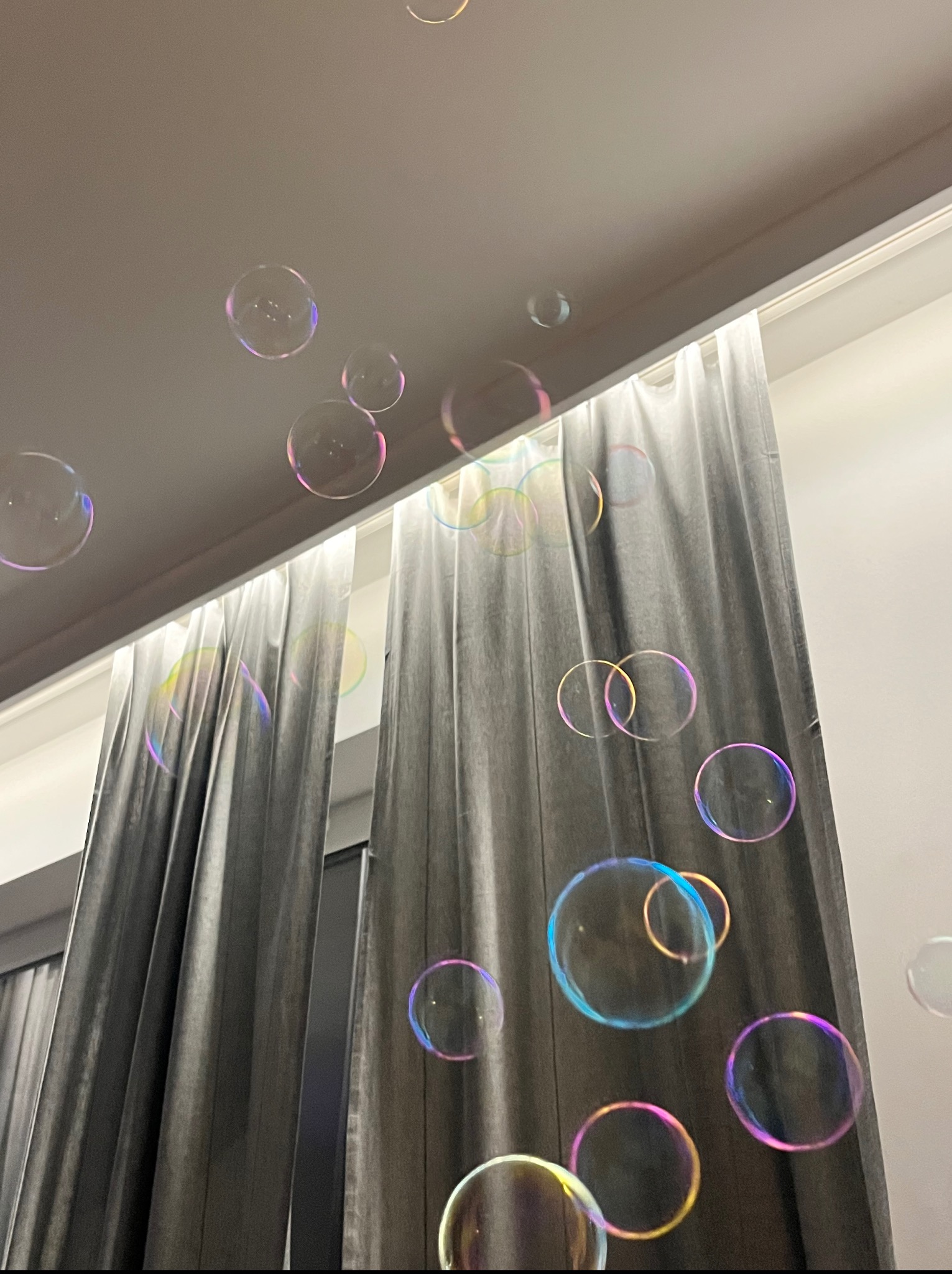 Lekkie, opalizujące bańki mydlane unoszą się w powietrzu na tle szarych zasłon i białego sufitu. Delikatne światło podkreśla ich okrągły kształt i barwne refleksy.