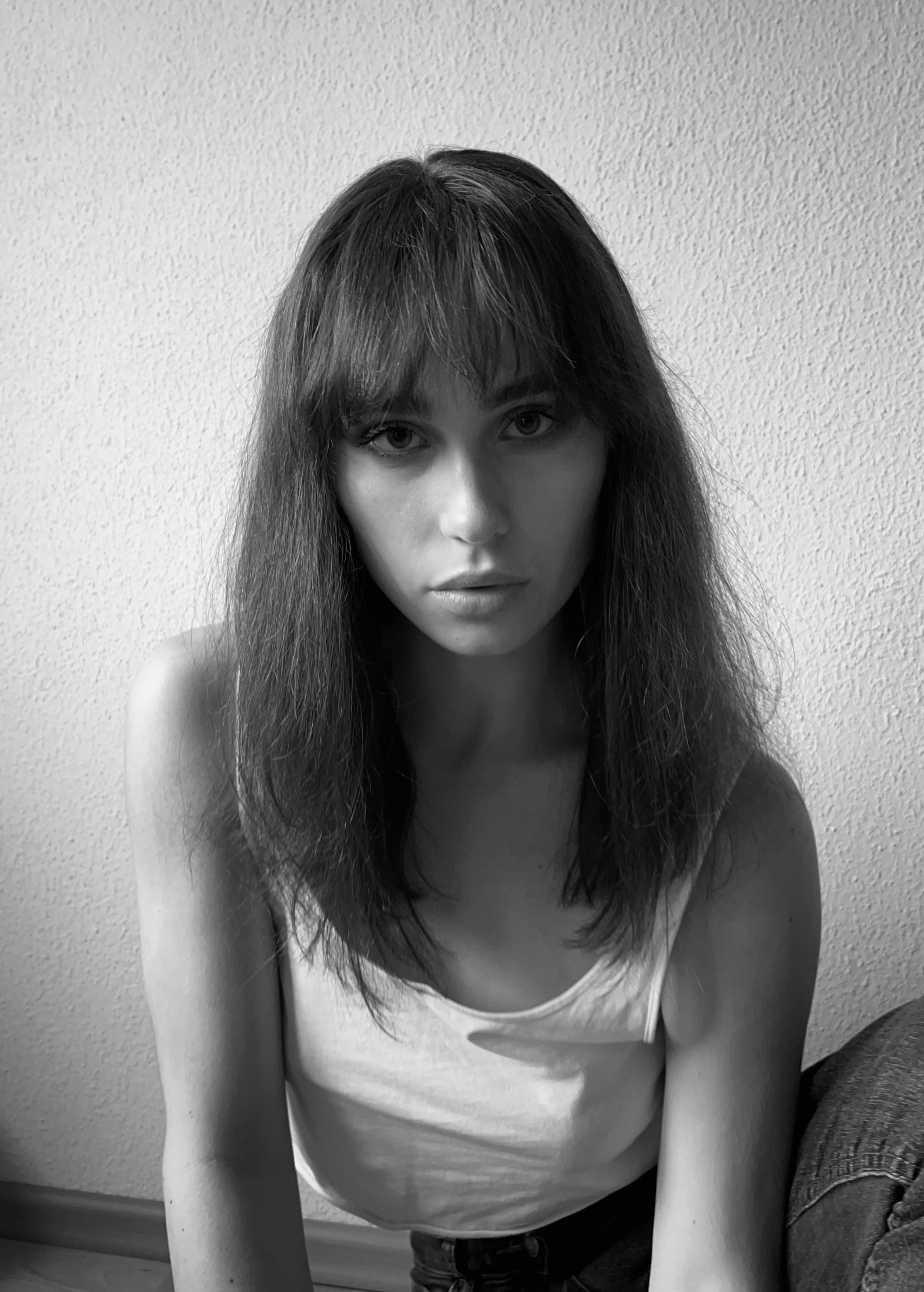 Monochromatyczny portret młodej kobiety z długimi włosami i grzywką, w białym topie, na tle jasnej ściany o wyraźnej fakturze. Ujęcie z dołu, podkreślające spojrzenie.