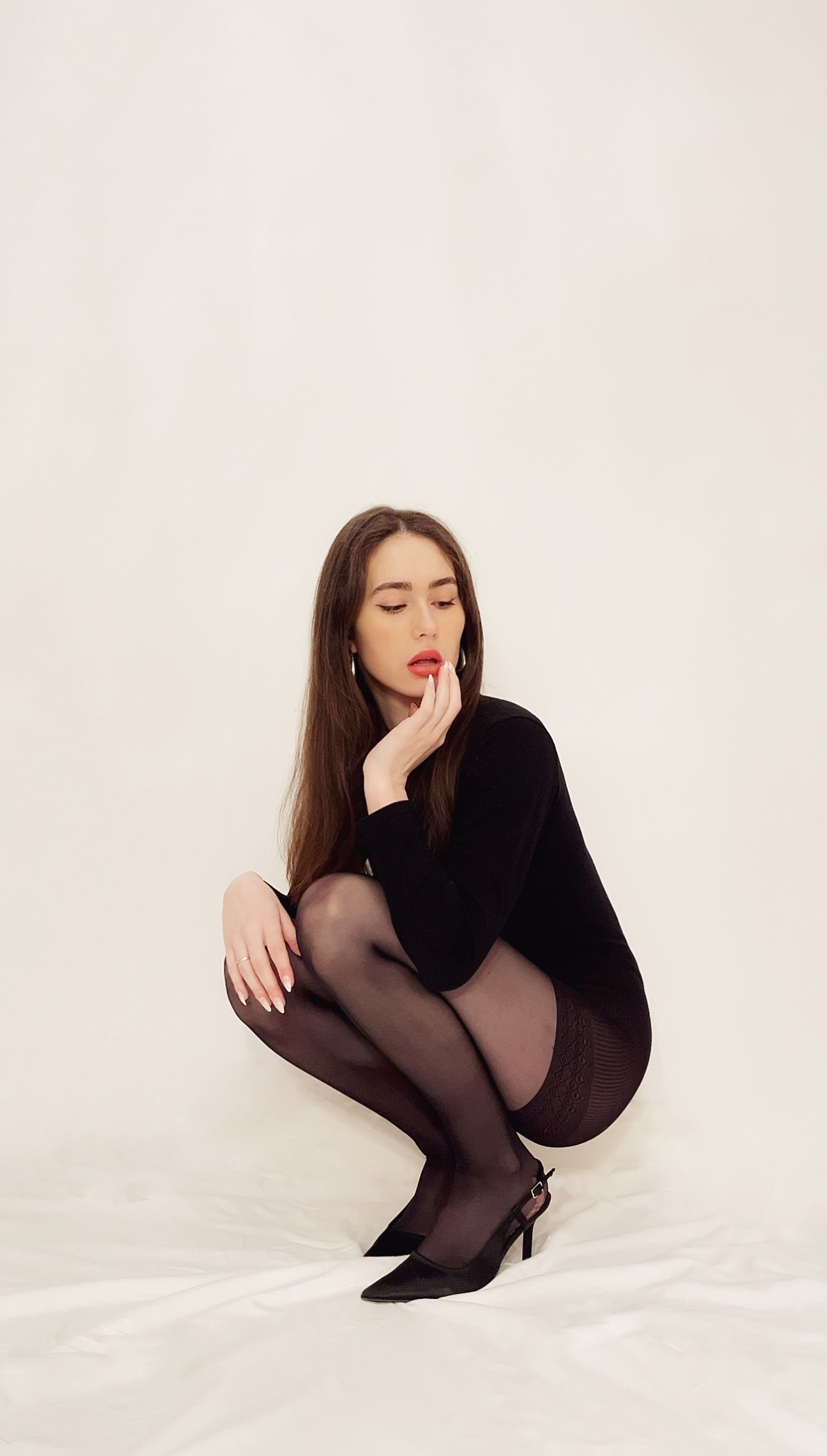 Portret kobiety w czarnym body, rajstopach i szpilkach, siedzącej na białym tle. Długie, brązowe włosy, czerwone usta, srebrne kolczyki. Delikatna poza i stonowane oświetlenie.