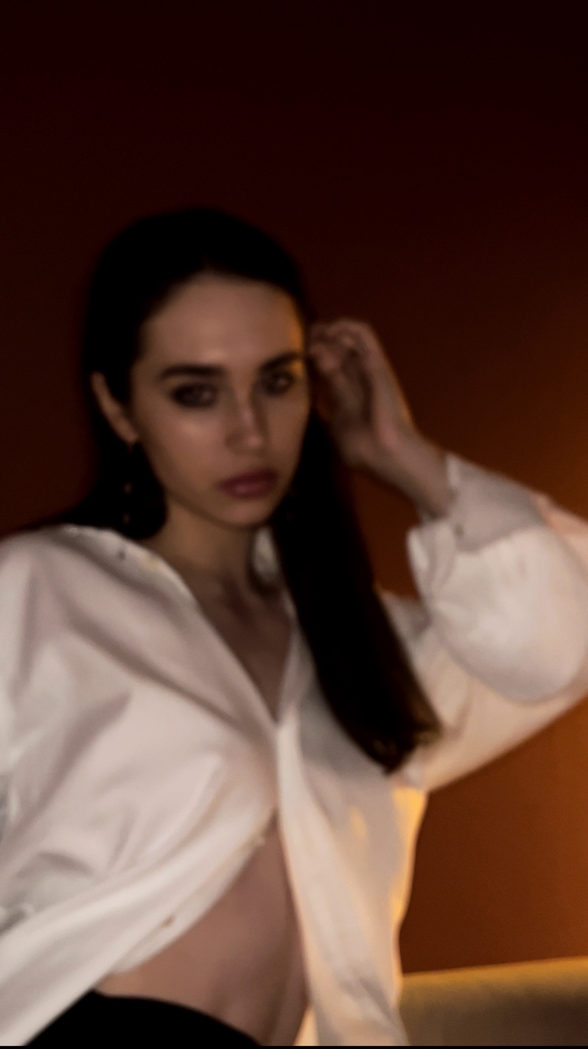 Portret kobiety z ciemnymi włosami w rozpiętej białej koszuli, w ciepłym oświetleniu. Wyraziste spojrzenie, delikatny makijaż, ręka uniesiona przy włosach.