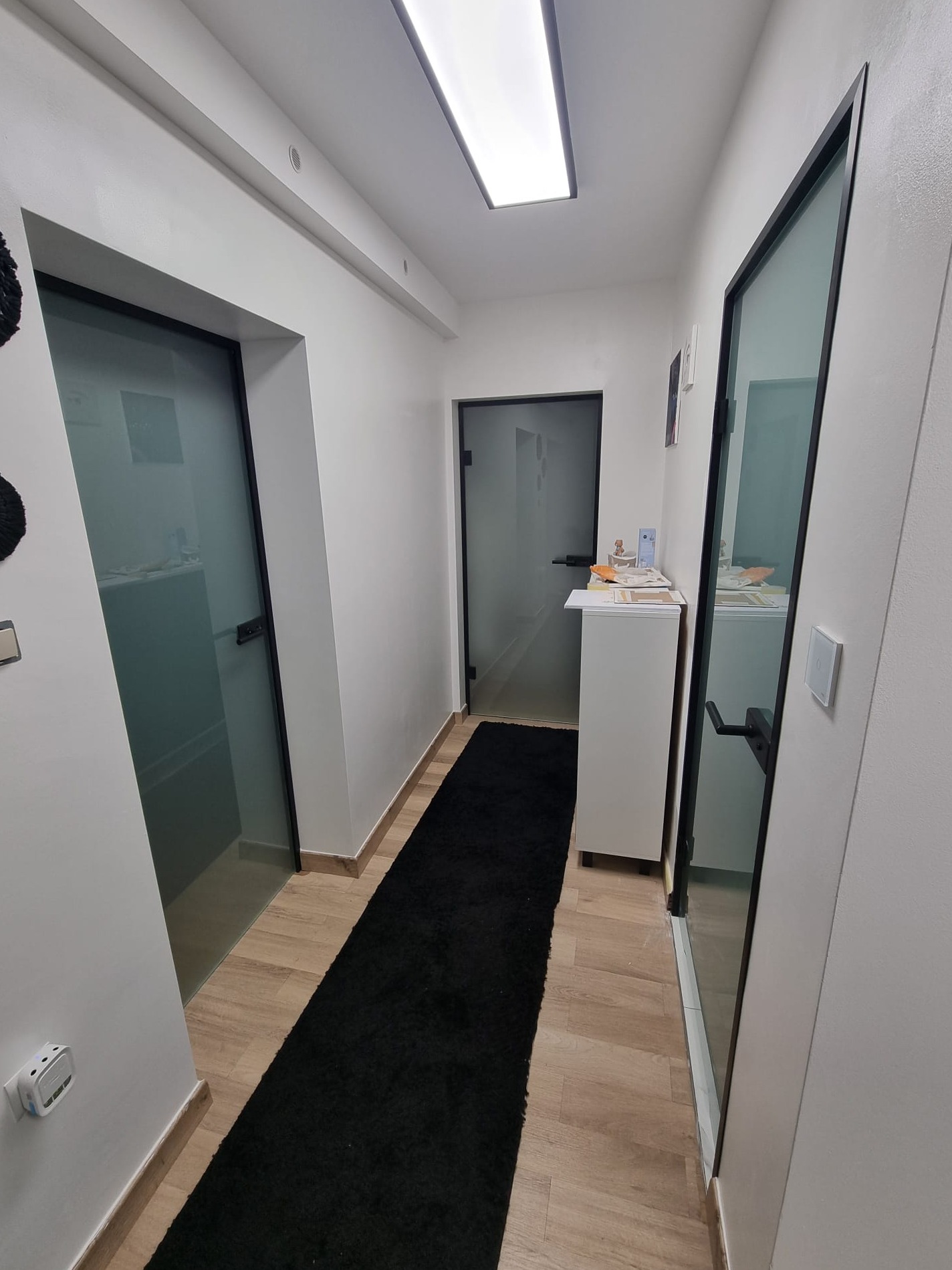 Korytarz biurowy z drewnianą podłogą, czarnym dywanem i szklanymi drzwiami z czarnymi ramami. Ściany pomalowane na biało, oświetlenie sufitowe.