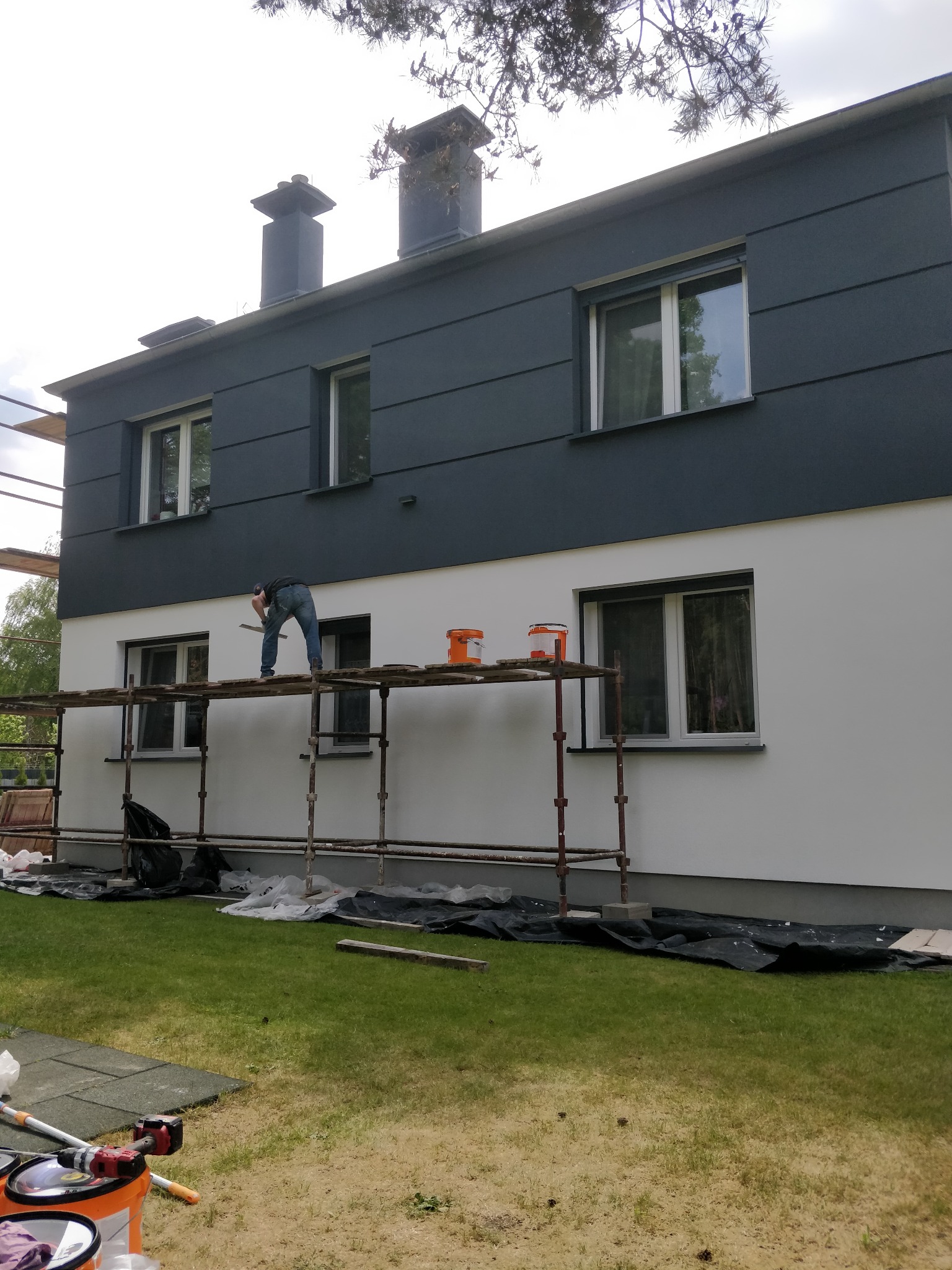 Dwukondygnacyjny dom z szarą górną częścią elewacji i białą dolną. Malarz na rusztowaniu wyrównuje ścianę. Widoczne wiaderka z farbą i narzędzia.