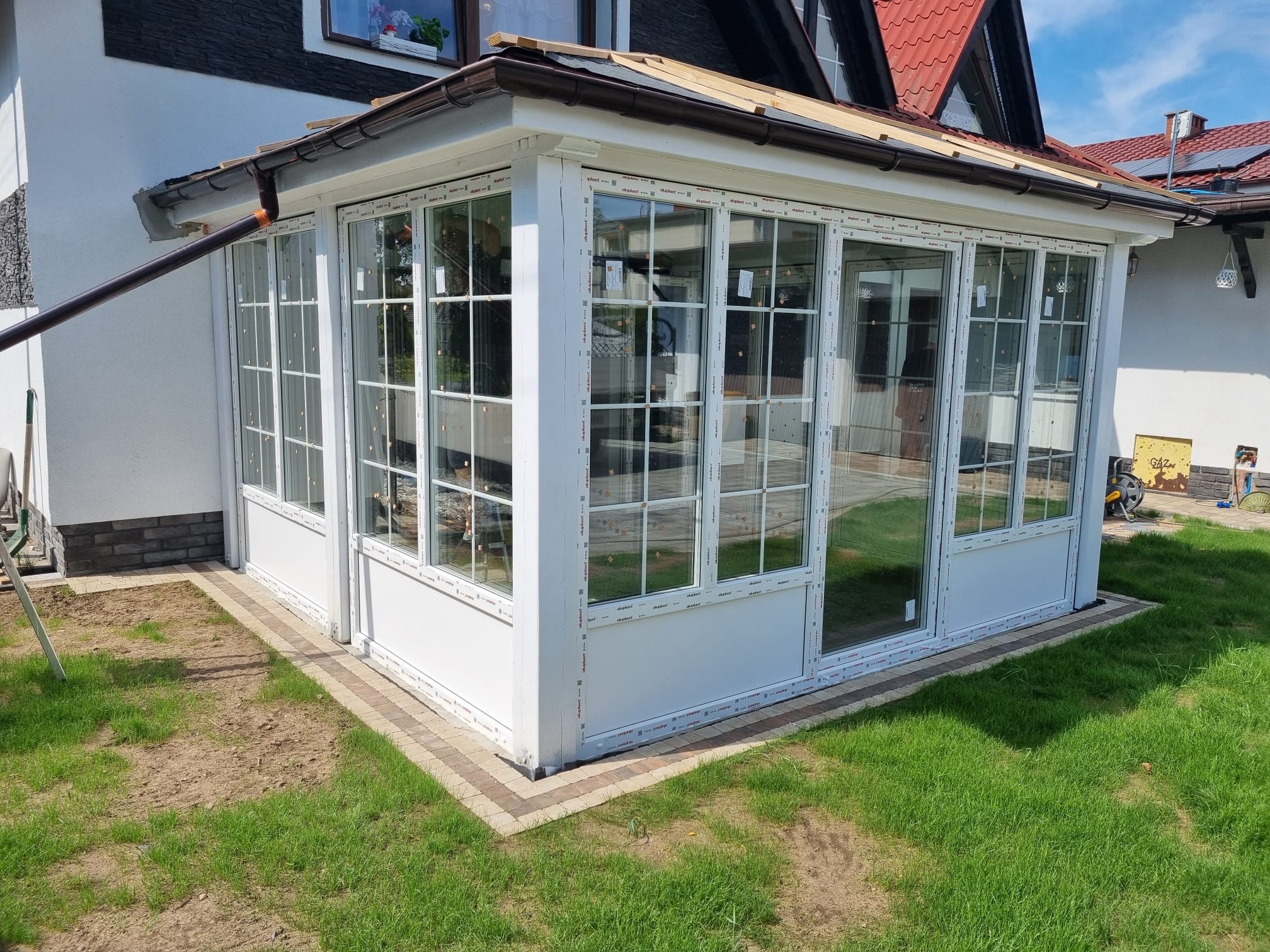 Nowoczesna, przeszklona weranda z białymi ramami okiennymi, wykończona ceglaną podmurówką, przylegająca do białego domu z ciemnym dachem. Widok z zewnątrz, trawnik.