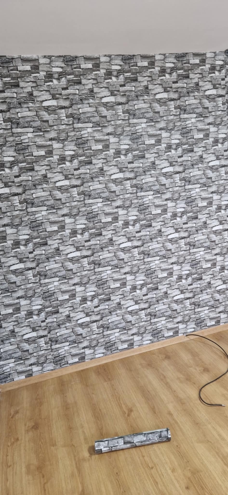 Ściana z tapetą imitującą kamień w odcieniach szarości, z fragmentem rolki tapety na podłodze. Widoczna drewniana podłoga i listwa przypodłogowa.
