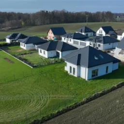 Osiedle domów jednorodzinnych z ciemnymi dachami, widok z lotu ptaka. Jeden dom w trakcie budowy, zabezpieczony siatką. Zielone trawniki i pola uprawne w tle.