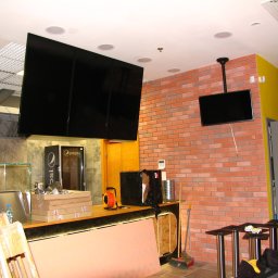 Pe-Jot Piotr Jurkowski - Montaż ekranów w restauracji: widoczne trzy duże monitory i jeden mniejszy na ceglanej ścianie. W tle lodówka Pepsi i elementy baru w trakcie instalacji.