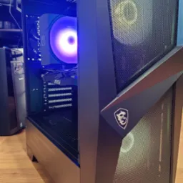 Komputer stacjonarny w czarnej obudowie MSI z podświetleniem LED, widoczny wentylator i karta graficzna. Sprzęt na drewnianej podłodze.