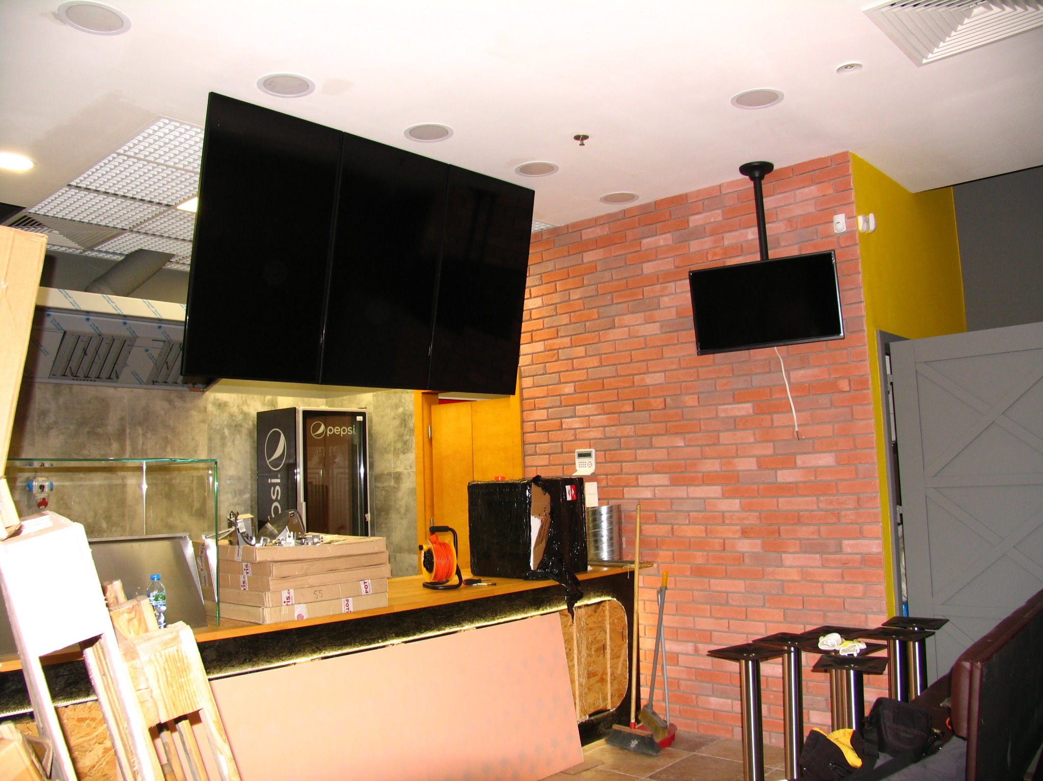 Montaż ekranów w restauracji: widoczne trzy duże monitory i jeden mniejszy na ceglanej ścianie. W tle lodówka Pepsi i elementy baru w trakcie instalacji.