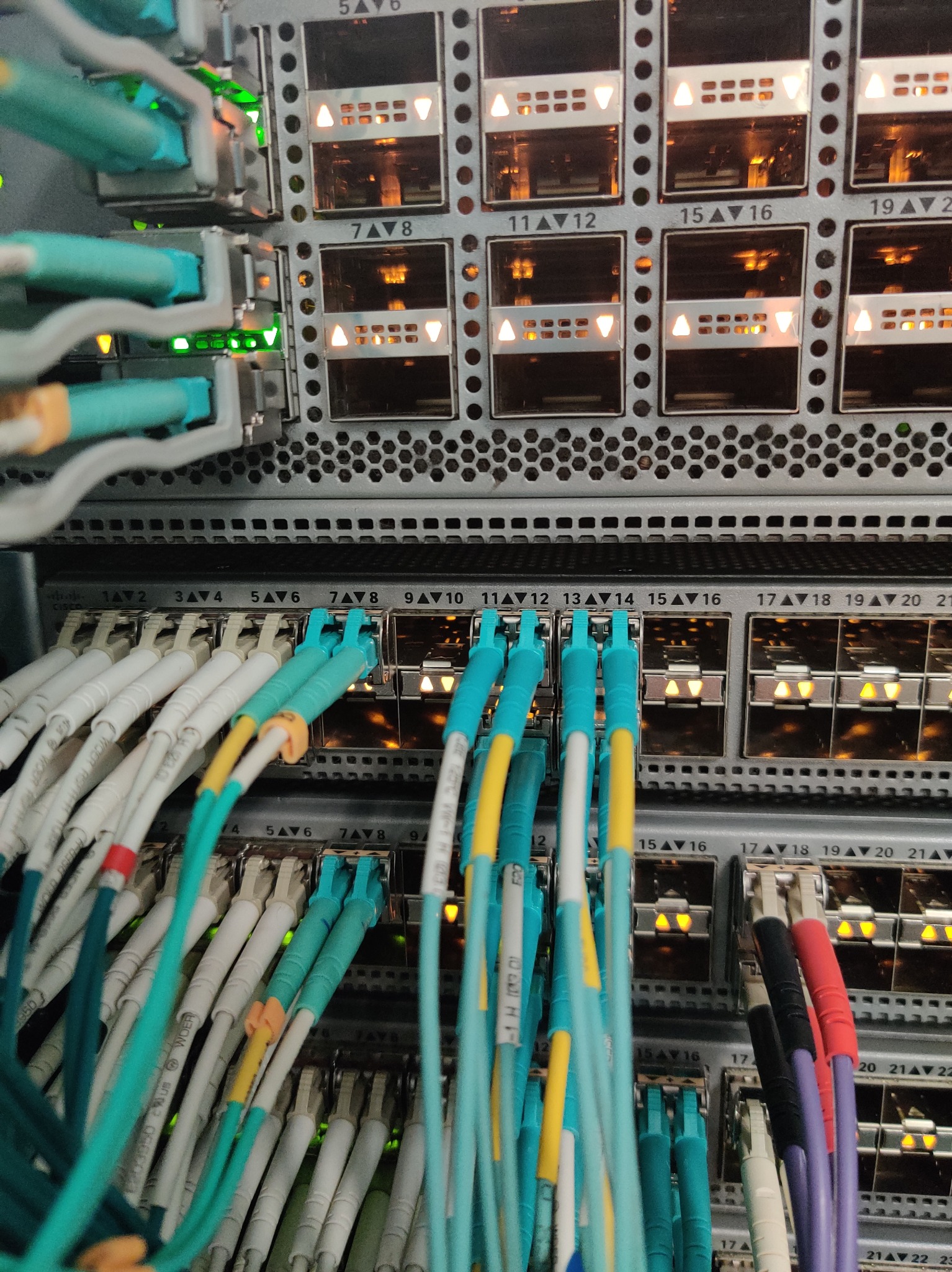 Szczegółowe ujęcie szafy rack z precyzyjnie ułożonymi kablami światłowodowymi oznaczonymi kolorowymi taśmami, w tle widoczne porty z aktywnymi diodami.