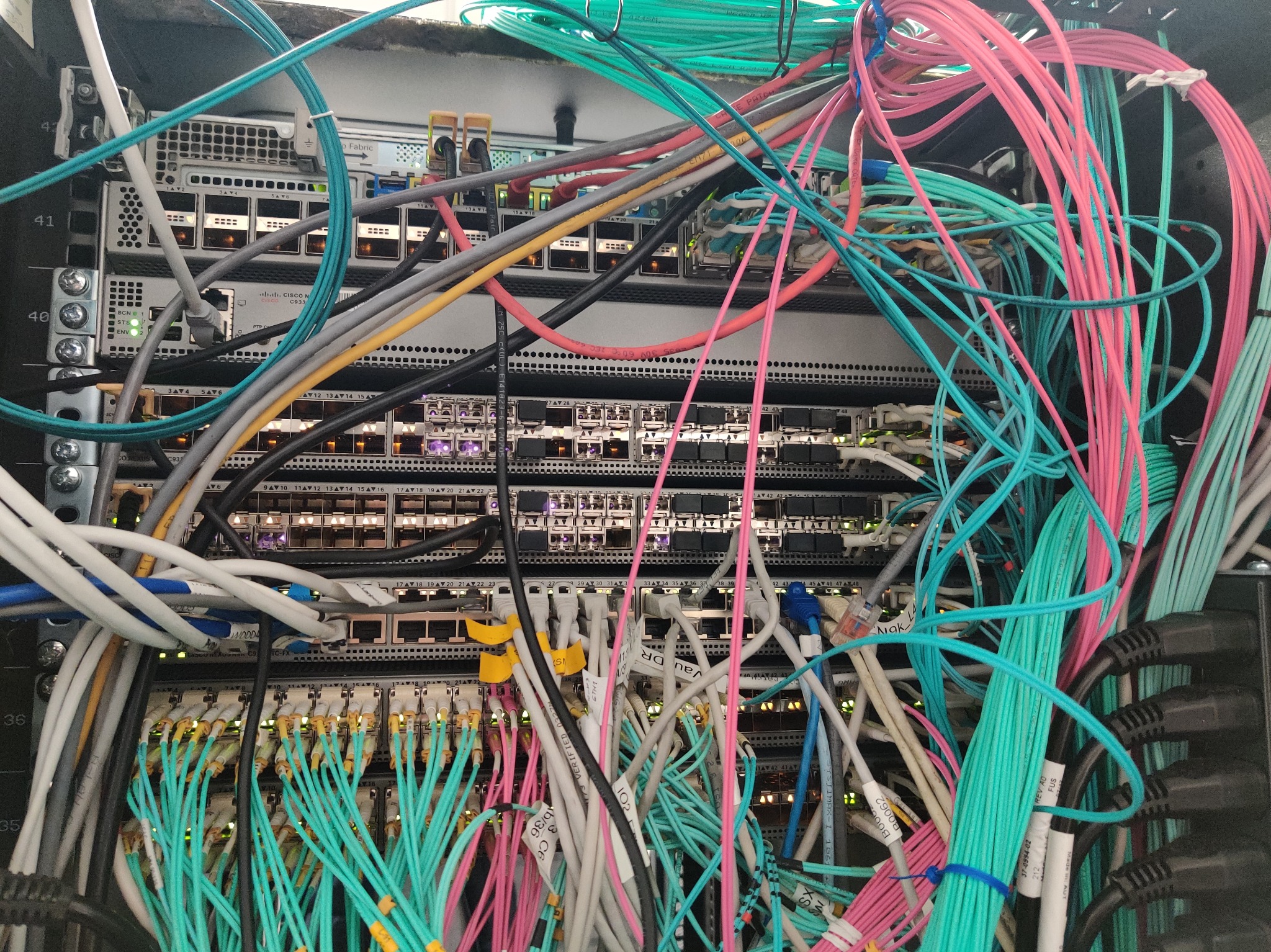Skomplikowana sieć kabli w szafie rack, z widocznymi przełącznikami Cisco i różnokolorowymi przewodami oznaczonymi etykietami, tworzącymi gęstą, techniczną plątaninę.