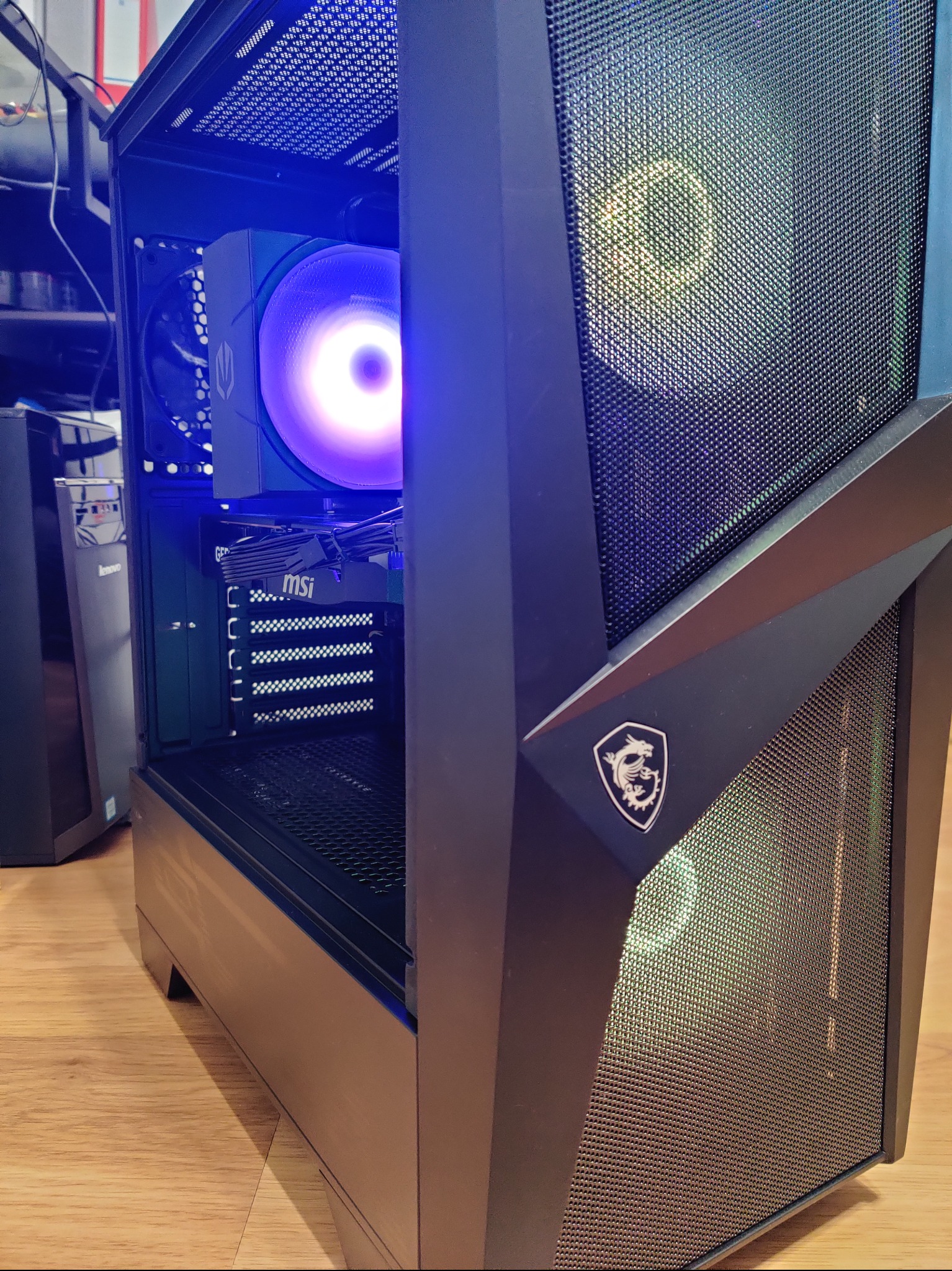 Komputer stacjonarny w czarnej obudowie MSI z podświetleniem LED, widoczny wentylator i karta graficzna. Sprzęt na drewnianej podłodze.