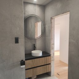 Custom Constructions - Nowoczesna łazienka z szarymi płytkami imitującymi beton, drewnianą szafką pod umywalkę z ryflowanym frontem, okrągłym lustrem i czarną armaturą. Widoczne niedokończone ościeże drzwiowe.
