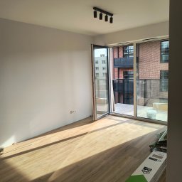Custom Constructions - Puste, jasne pomieszczenie z nową podłogą panelową w odcieniu jasnego drewna, białymi ścianami i czarną lampą sufitową. Duże drzwi balkonowe prowadzą na balkon z widokiem na ceglany budynek. W tle...
