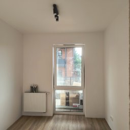 Custom Constructions - Pusty pokój po remoncie z białymi ścianami, jasną podłogą panelową i nowoczesnym oświetleniem. Na grzejniku tuby z silikonem, za oknem opakowania Cersanit.