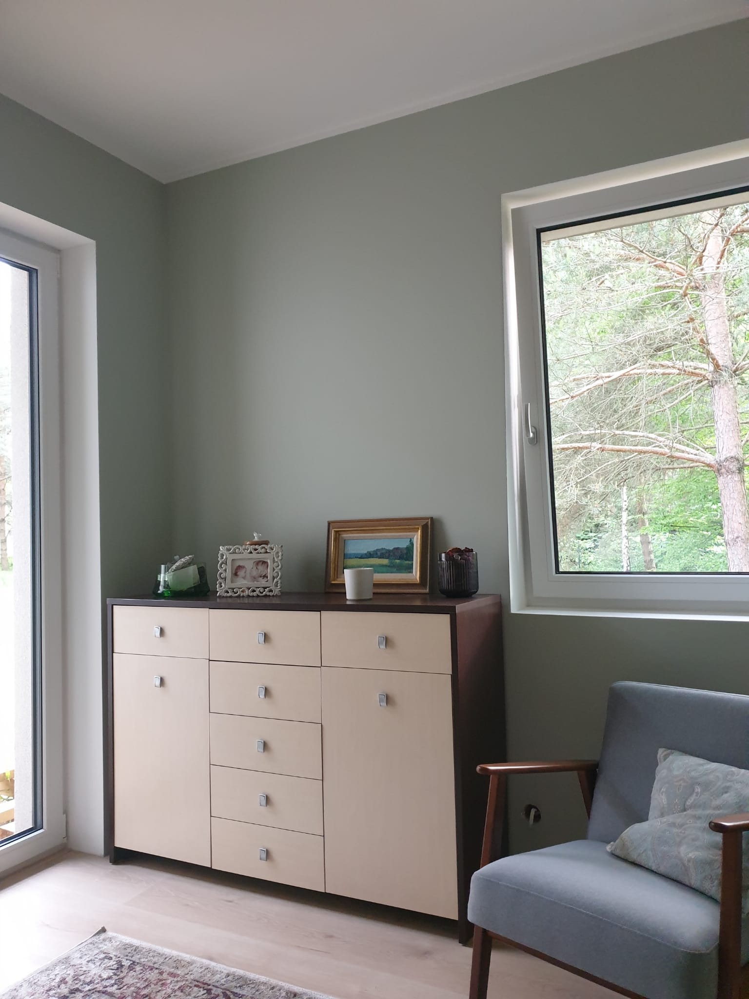 Pomalowane na szaro wnętrze pokoju z komodą, oknem z widokiem na drzewa i fotelem w stylu retro. Ściany gładkie, bez widocznych niedoskonałości.