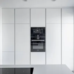 Minimalistyczna kuchnia z zabudową w bieli, z wbudowanym piekarnikiem Bosch i blatem z czarną płytą indukcyjną. Nowoczesny design z oknem po prawej stronie.