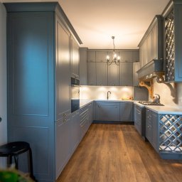 KAPI Szwarckopf - Meble Na Wymiar - Szara kuchnia w stylu klasycznym z drewnianą podłogą. Uwagę przykuwają ozdobne szafki wiszące z ażurowymi frontami i fornirowany okap nad kuchenką gazową.