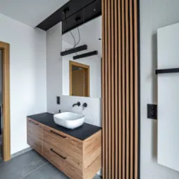 Nowoczesna łazienka z drewnianą szafką pod umywalką, białymi cegiełkami na ścianie i dekoracyjnymi drewnianymi lamelami. Minimalistyczne oświetlenie.