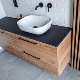 Nowoczesna szafka łazienkowa z drewna z czarnym blatem i białą umywalką nablatową. Minimalistyczna armatura w kolorze czarnym. Na ścianie biała cegła.