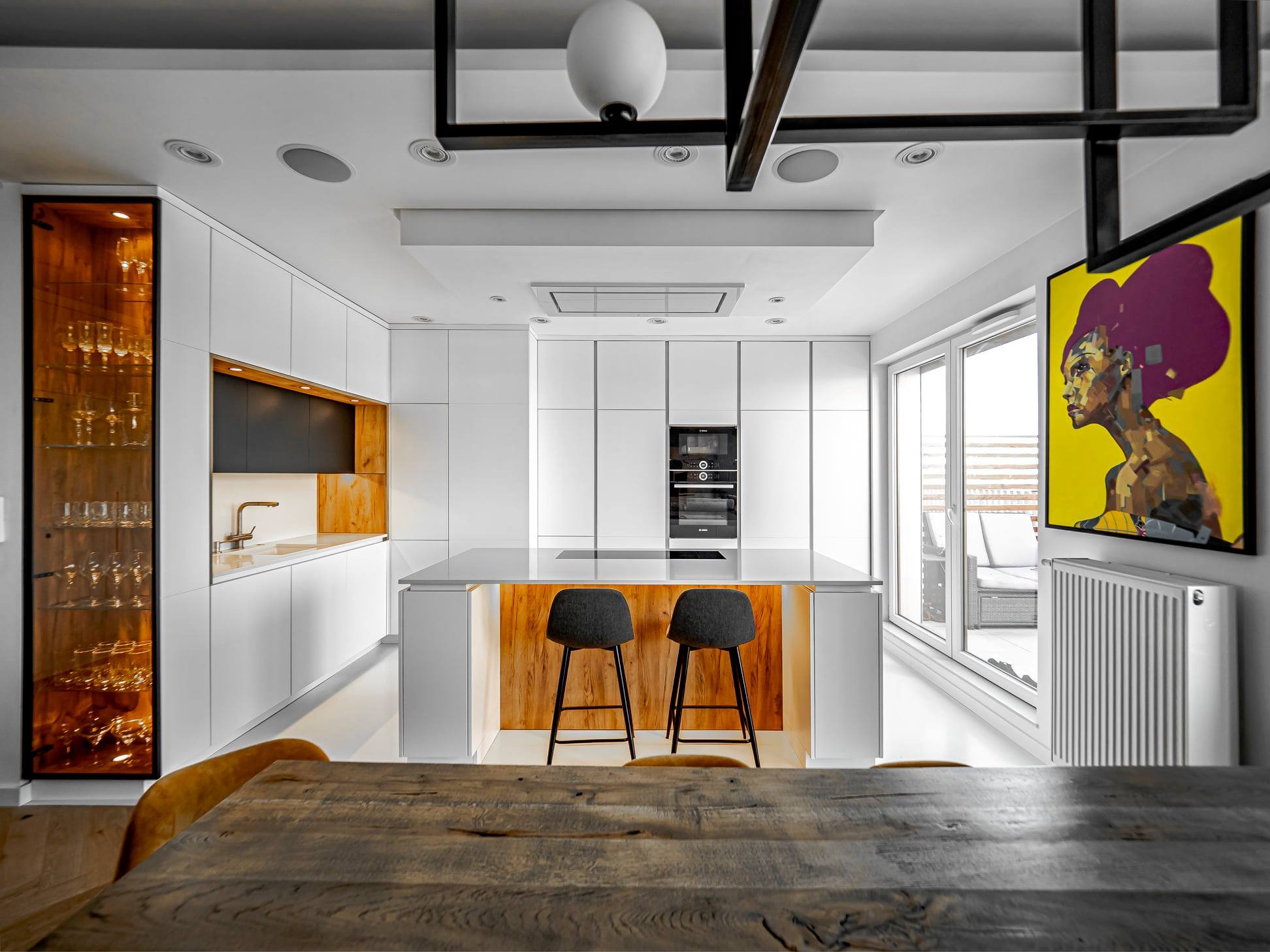 Nowoczesna kuchnia z wyspą i drewnianymi akcentami. Białe szafki, czarne blaty. Dwa krzesła barowe przy wyspie. Na ścianie żółty obraz.