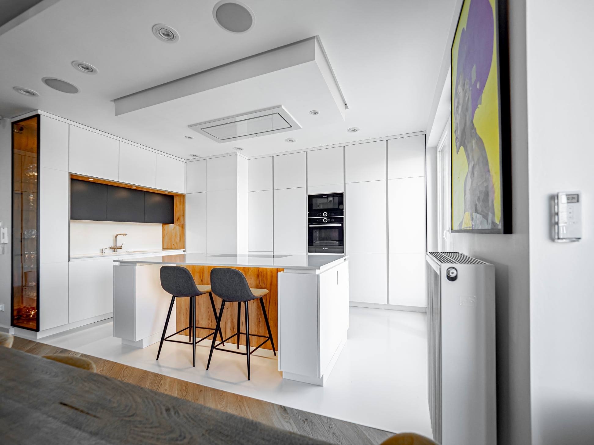 Nowoczesna kuchnia z wyspą i hokerami, białe fronty szafek, drewniane akcenty, oświetlenie sufitowe. Minimalistyczny design, obraz na ścianie.