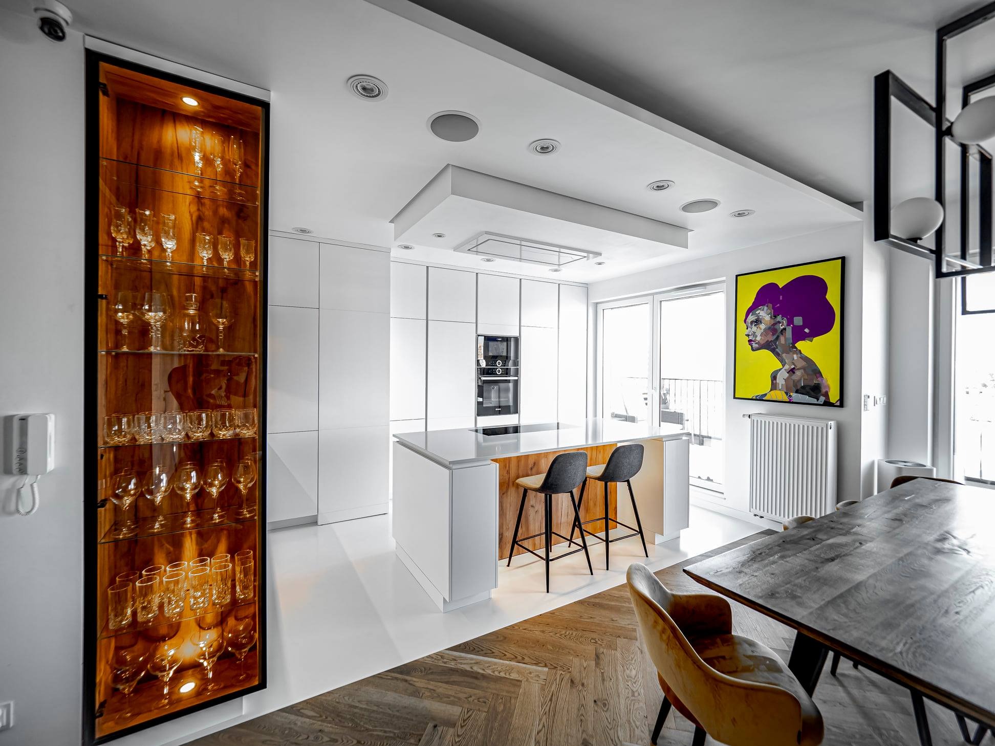 Nowoczesna, jasna kuchnia z wyspą i drewnianą witryną na szkło. Minimalistyczne białe szafki, parkiet w jodełkę i żółty obraz dodają charakteru wnętrzu.