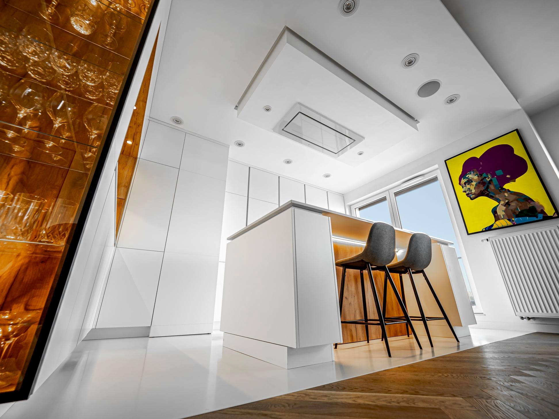 Nowoczesna, biała kuchnia z wyspą i szklaną witryną na alkohole. Uwagę przyciąga obraz na ścianie i drewniana podłoga ułożona w jodełkę. Nietypowy kadr z dołu.