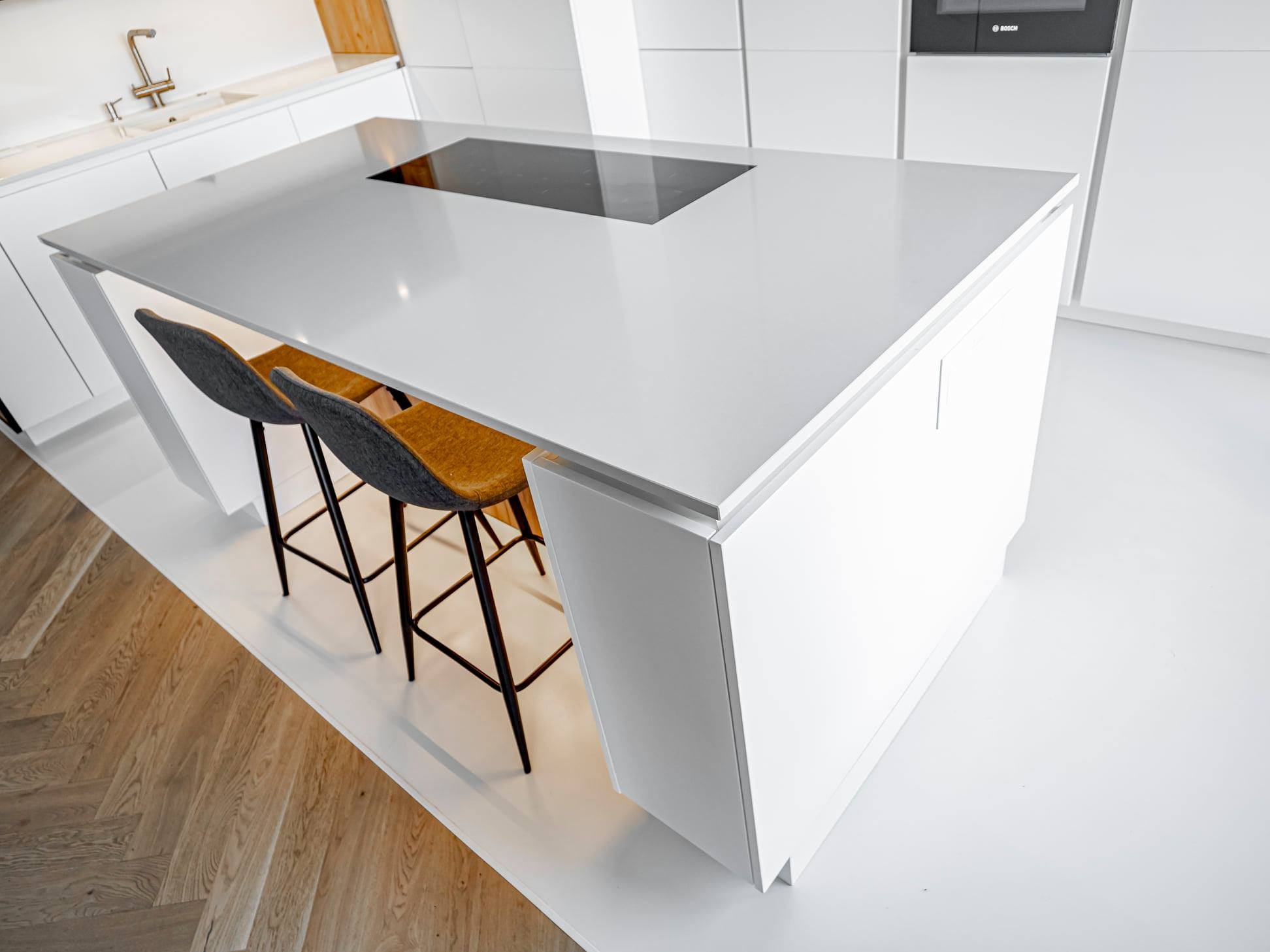 Minimalistyczna, biała wyspa kuchenna z płytą indukcyjną i dwoma krzesłami barowymi na tle jasnej podłogi i zabudowy kuchennej.