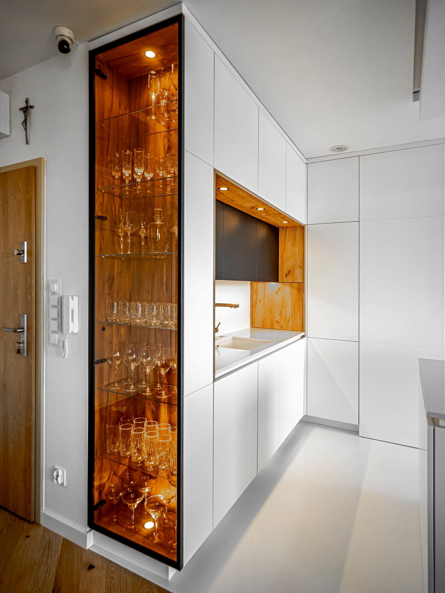 Nowoczesna kuchnia z białymi szafkami i przeszkloną witryną z podświetleniem, eksponującą kolekcję kieliszków. Minimalistyczny design, ciepłe oświetlenie.