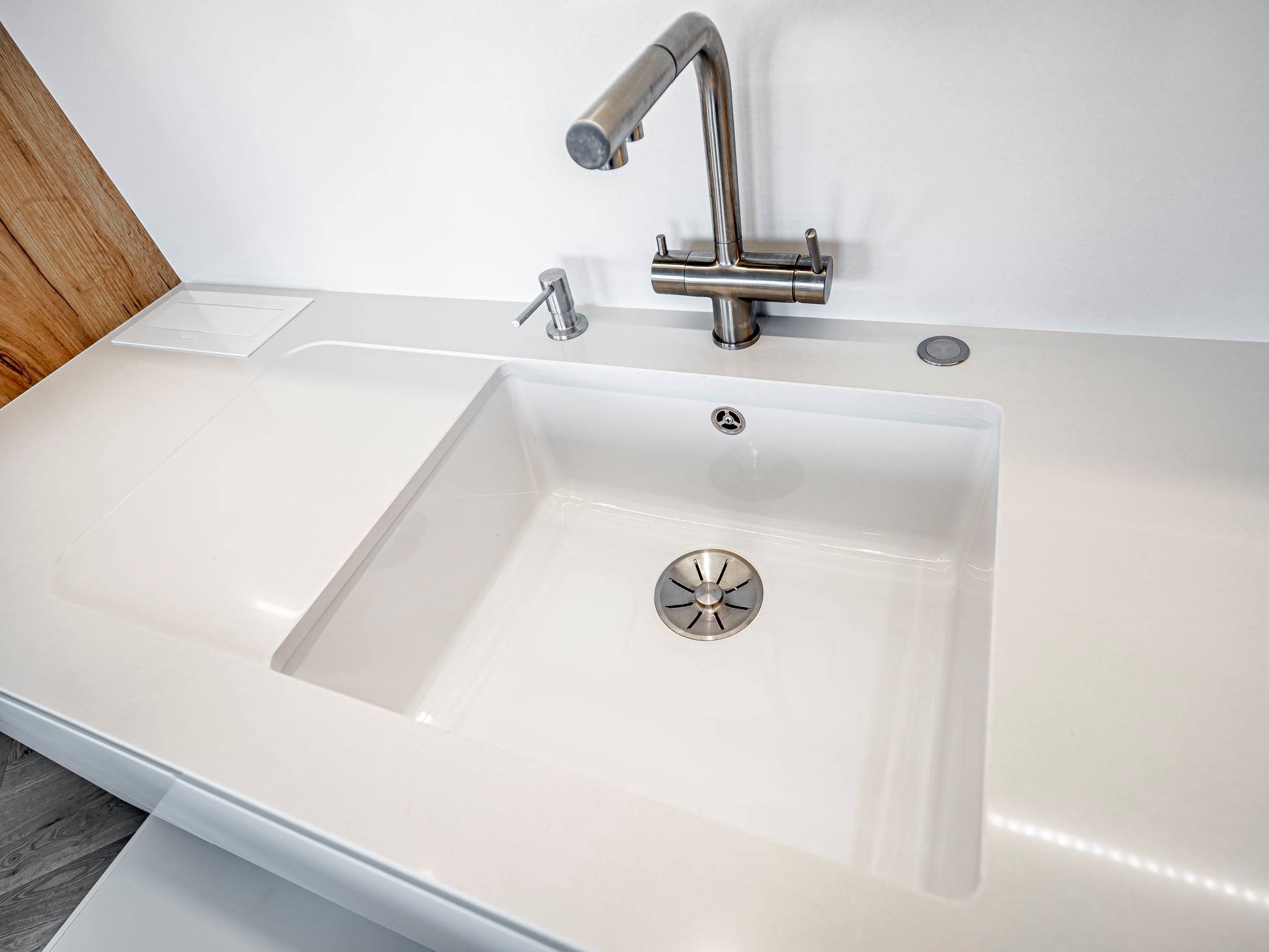 Biały, nowoczesny zlew kuchenny z chromowaną baterią i dozownikiem mydła, wkomponowany w blat. Widoczny fragment drewnianej zabudowy. Minimalistyczny design.