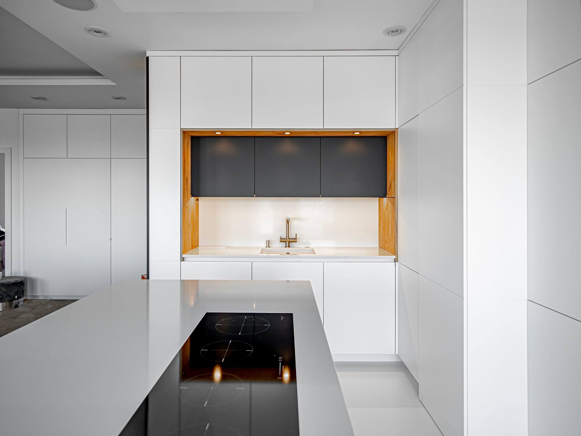 Nowoczesna kuchnia z białymi szafkami i wyspą z płytą indukcyjną. Minimalistyczny design z drewnianymi akcentami i oświetleniem LED. Jasne wnętrze.