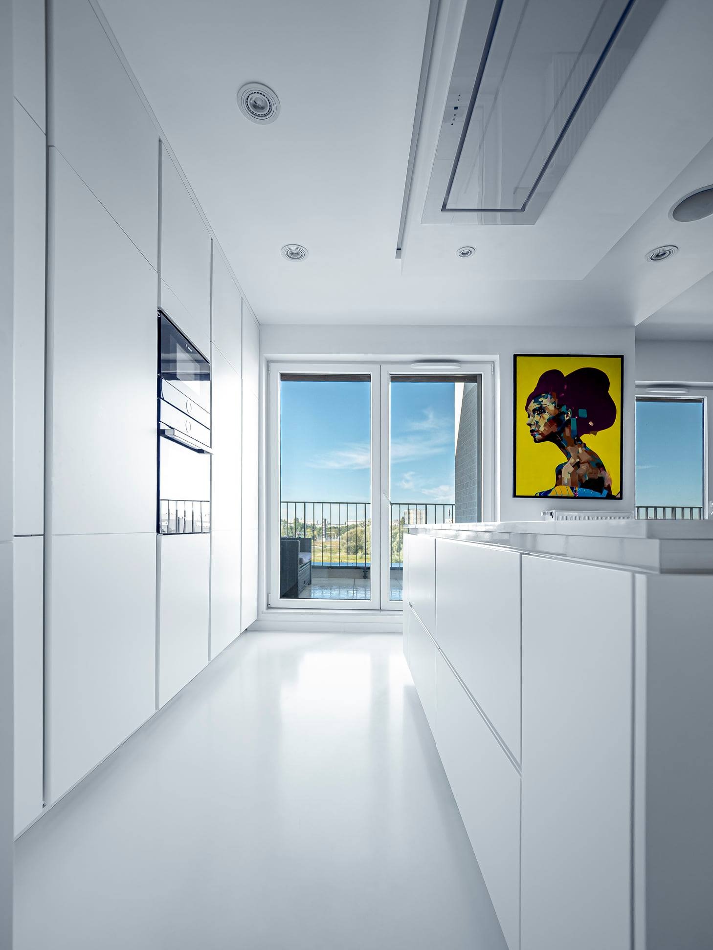 Nowoczesna, minimalistyczna kuchnia w bieli z wyjściem na balkon. Na ścianie kolorowy obraz. Oświetlenie sufitowe punktowe i okap w zabudowie.