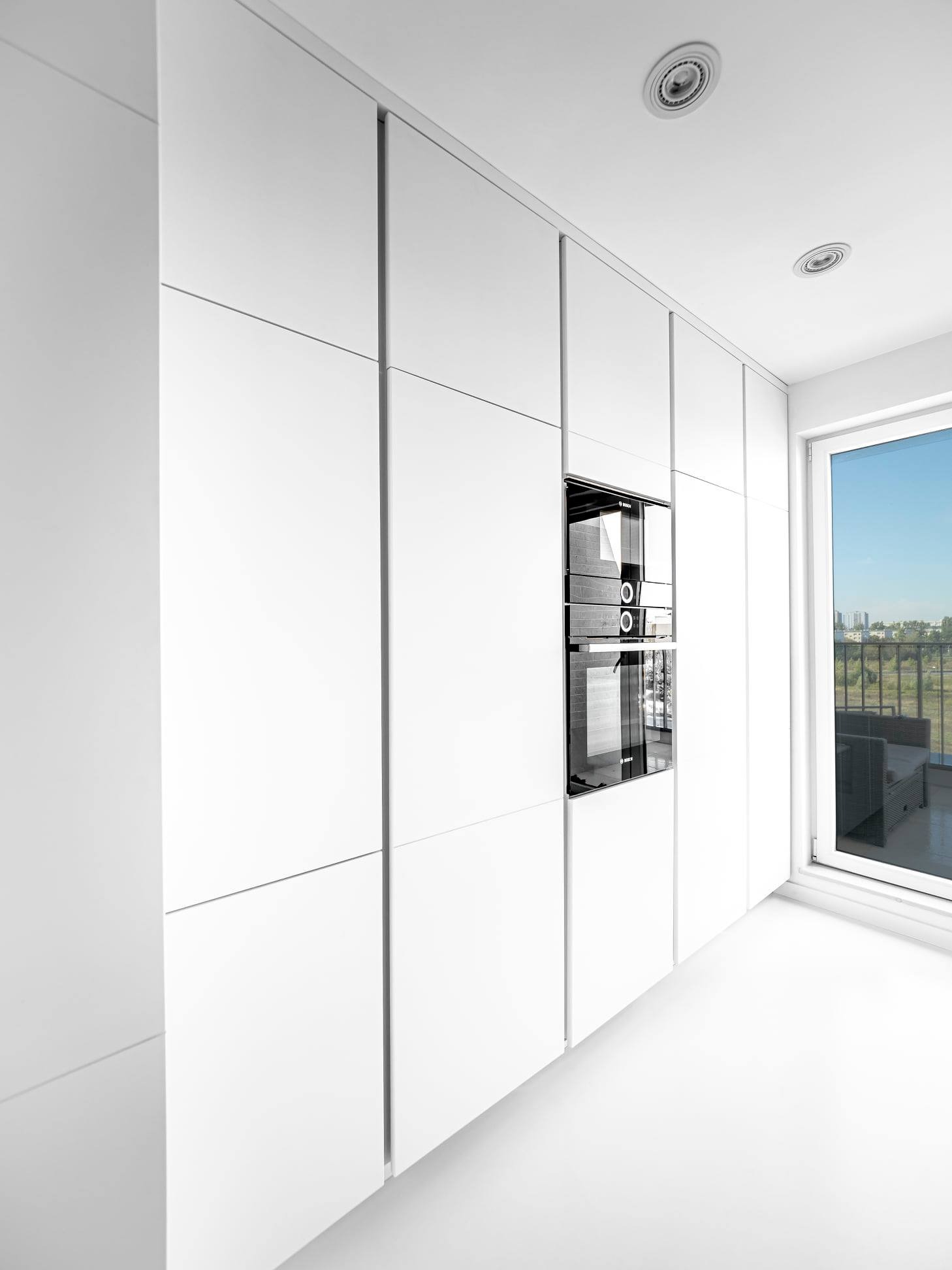 Minimalistyczna, biała kuchnia na wymiar z wbudowanym piekarnikiem i dużym oknem z widokiem na miasto. Nowoczesny design, czyste linie, eleganckie wykończenie.