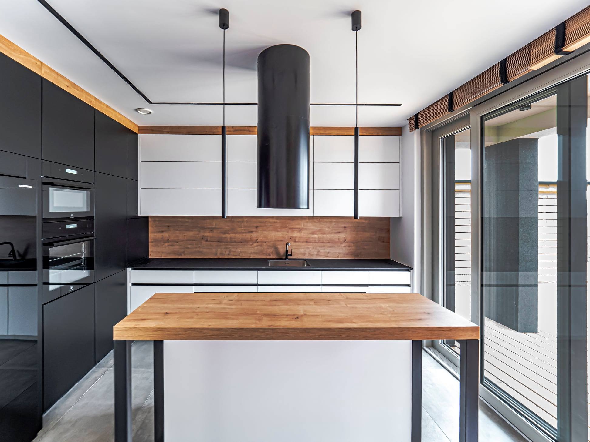 Nowoczesna kuchnia z wyspą: czarne szafki, białe górne szafki, drewniany blat i okap cylindryczny. Duże okno przesuwne z żaluzjami drewnianymi.