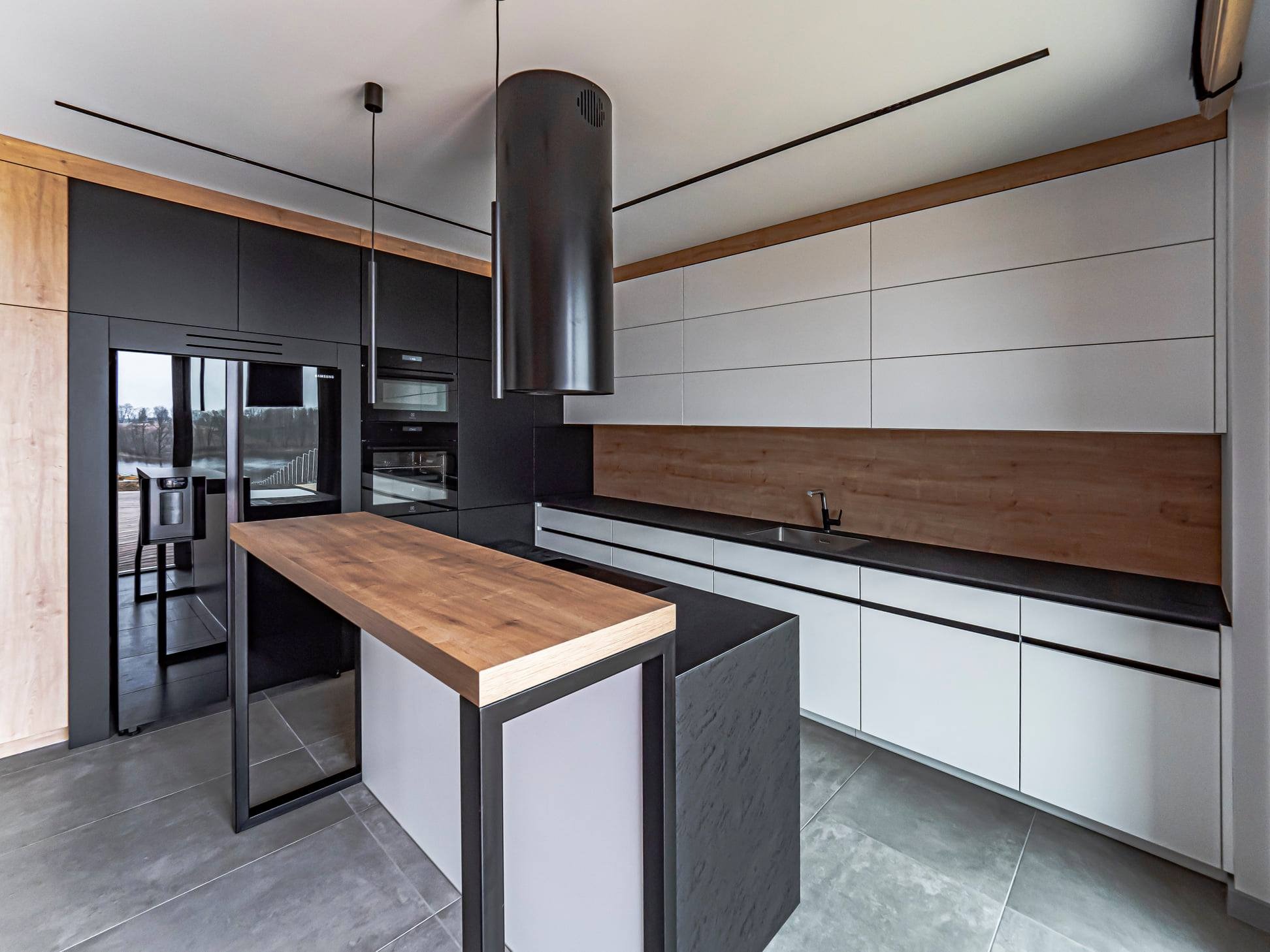 Nowoczesna kuchnia z wyspą: czarne i białe szafki, drewniane akcenty, minimalistyczna lampa nad wyspą. Szare płytki podłogowe, widok na zieleń za oknem.