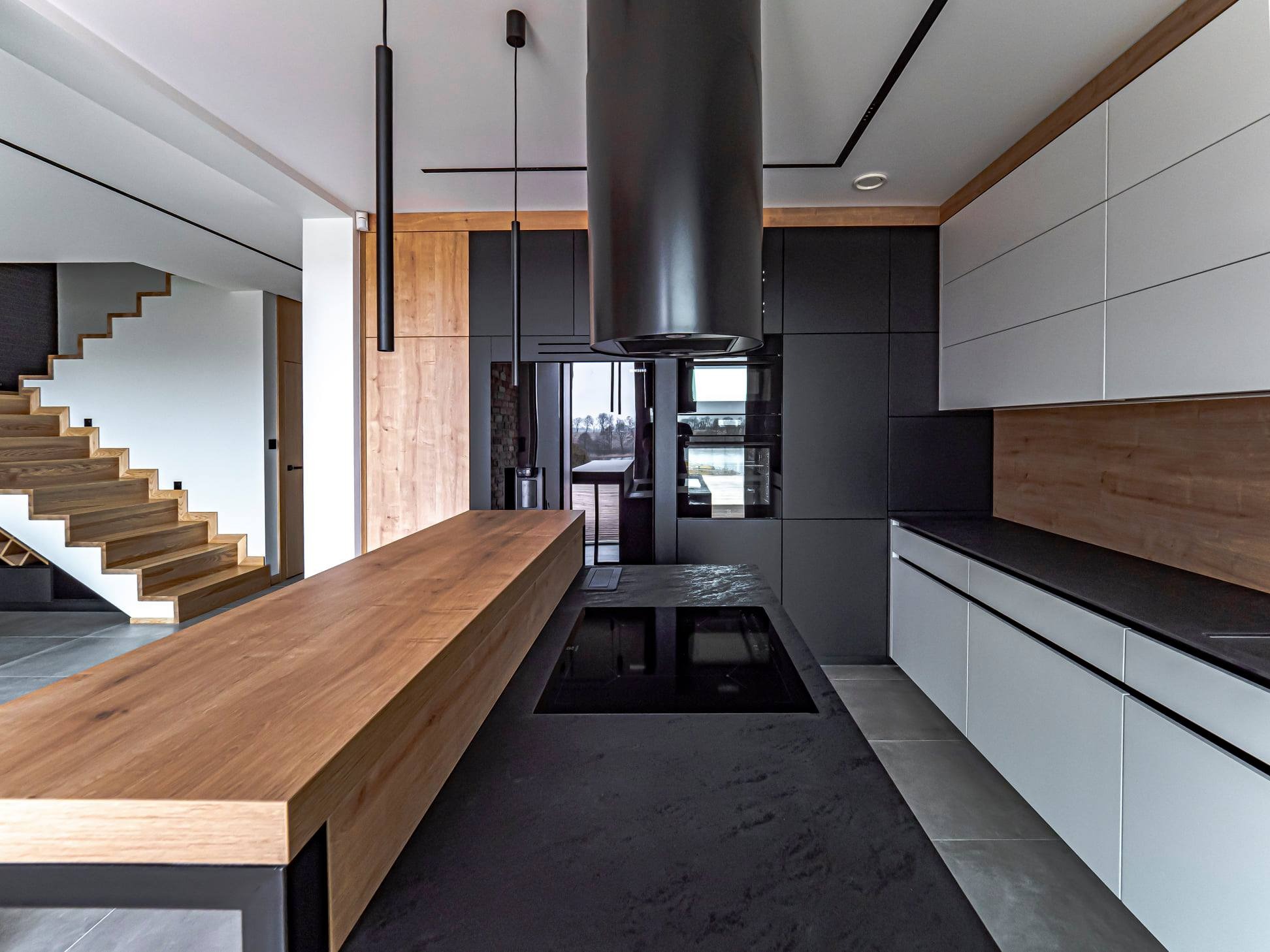 Nowoczesna kuchnia: czarne szafki, drewniany blat wyspy, minimalistyczny okap i schody z drewna. Kontrast materiałów i kolorów, eleganckie oświetlenie.
