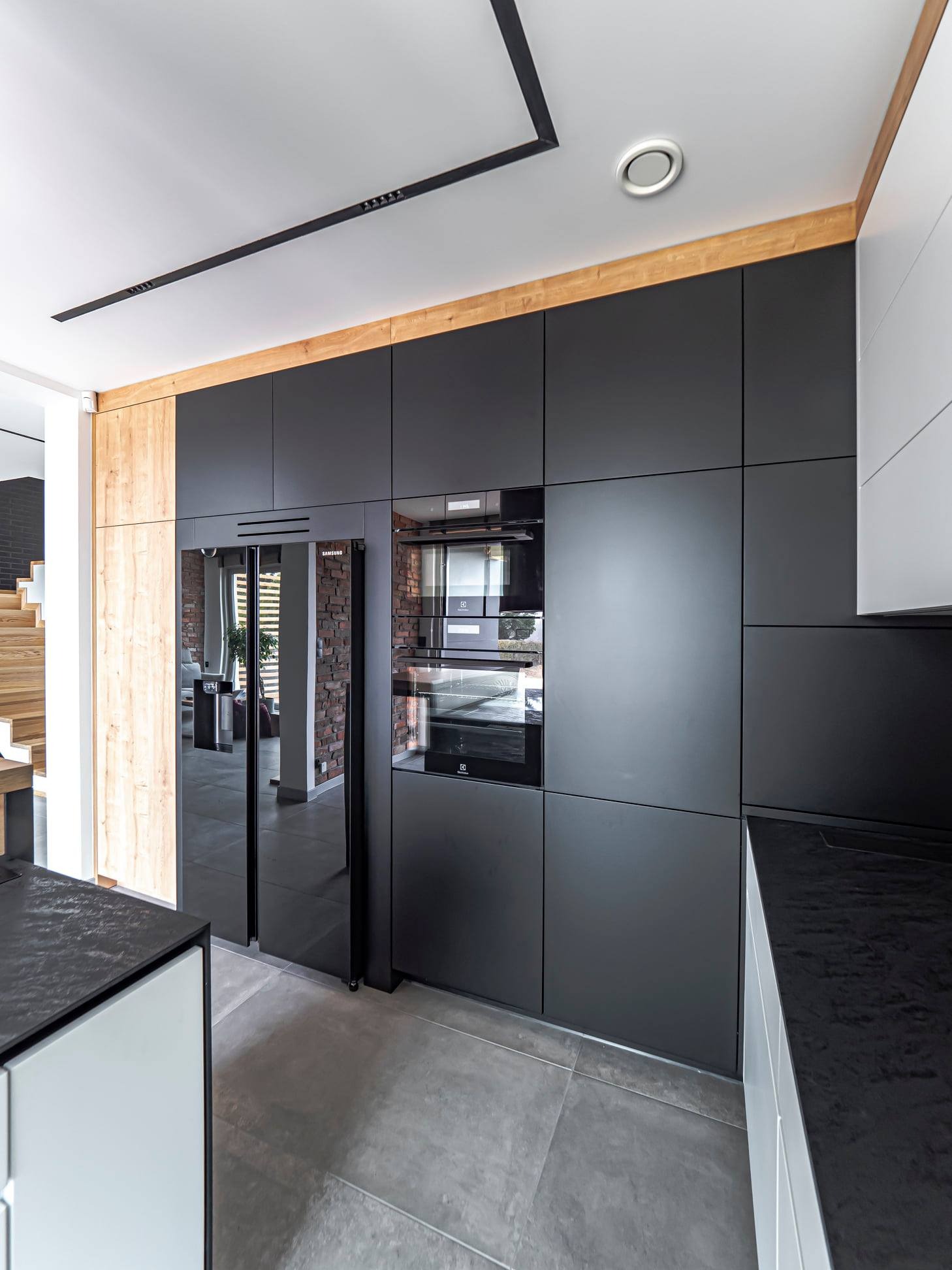 Nowoczesna kuchnia z czarnymi, matowymi frontami, drewnianymi akcentami i sprzętem AGD w zabudowie. Widoczna lodówka Samsung z czarnymi drzwiami.