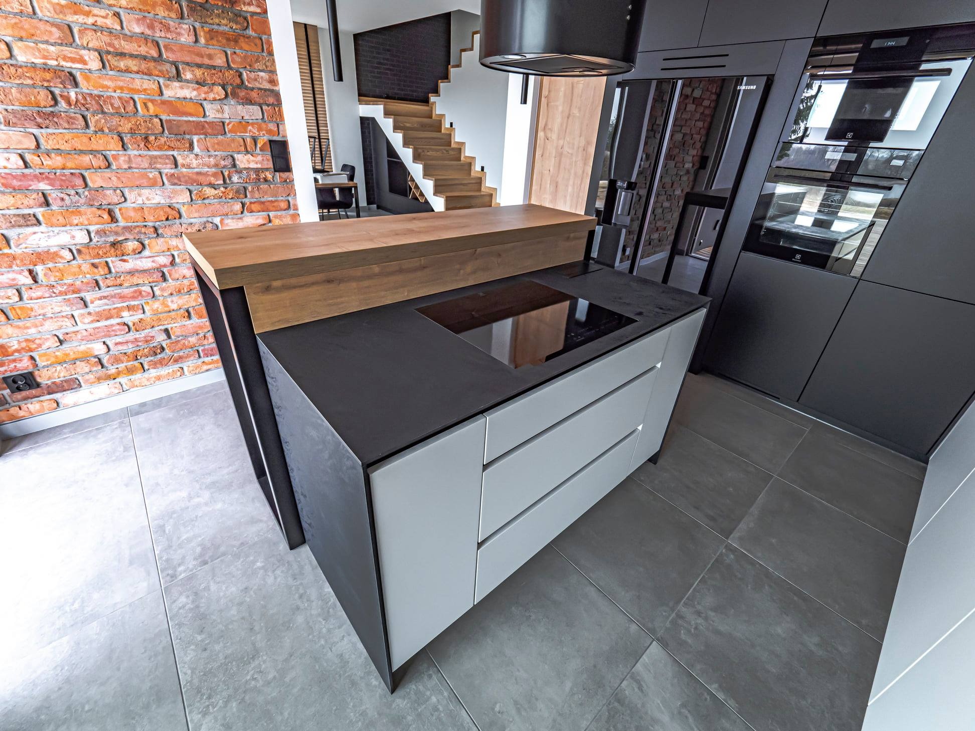 Wyspa kuchenna z płytą indukcyjną i drewnianym blatem na tle ceglanej ściany oraz schodów. Nowoczesne, czarne szafki kuchenne w zabudowie.