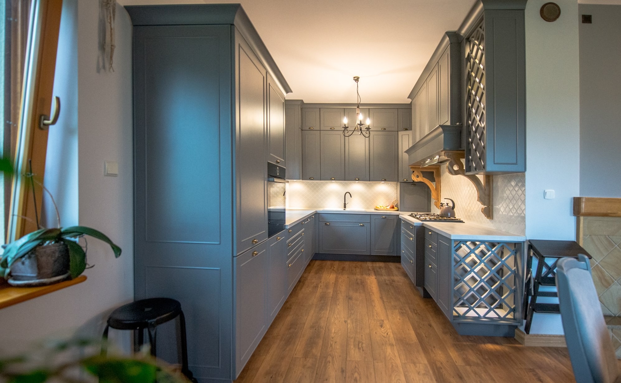 Szara kuchnia w stylu klasycznym z drewnianą podłogą. Uwagę przykuwają ozdobne szafki wiszące z ażurowymi frontami i fornirowany okap nad kuchenką gazową.