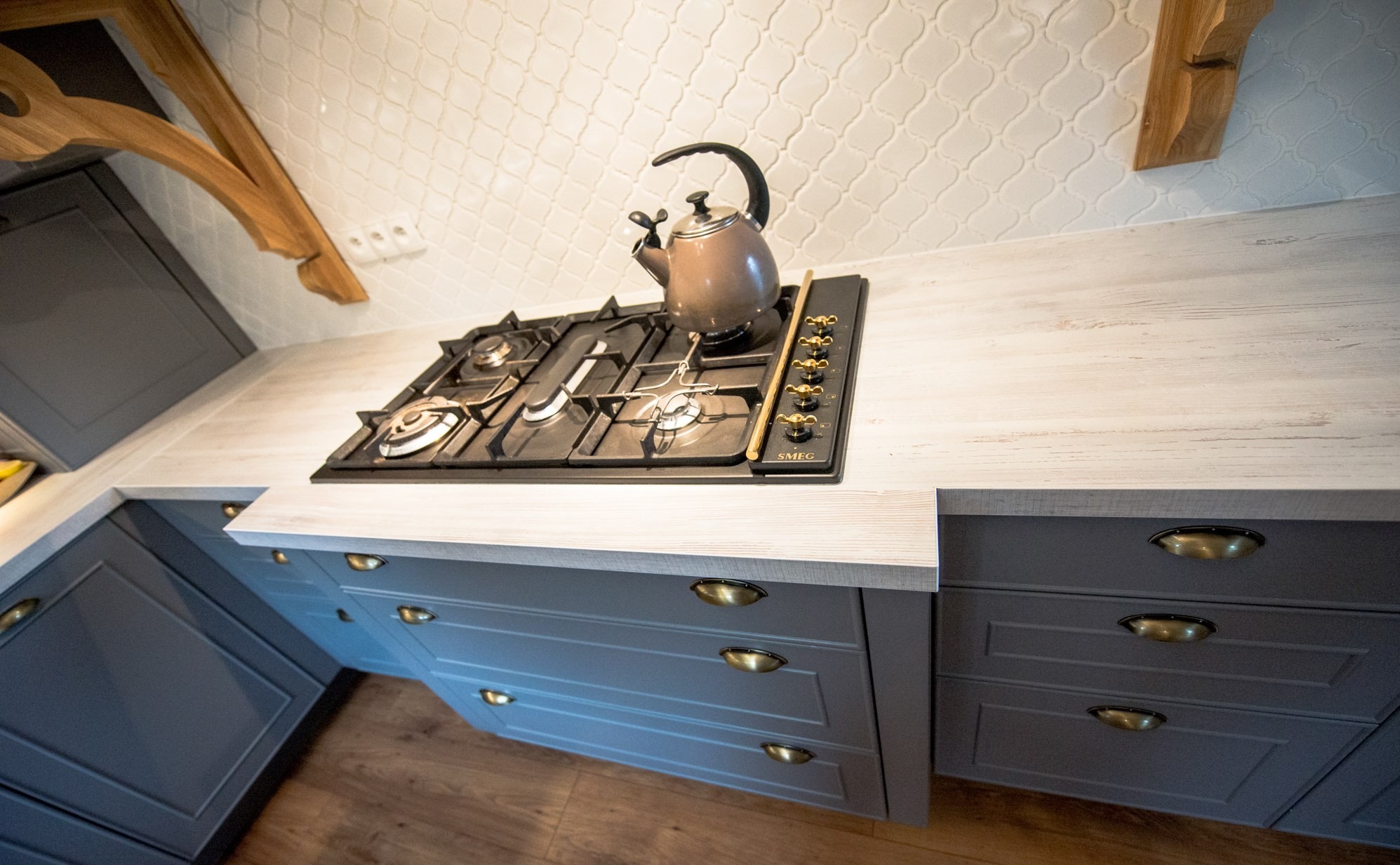 Szare szafki kuchenne z blatem imitującym drewno, płyta gazowa SMEG i czajnik na tle białych płytek w marokańskim stylu. Ujęcie z dołu, podkreślające detale.