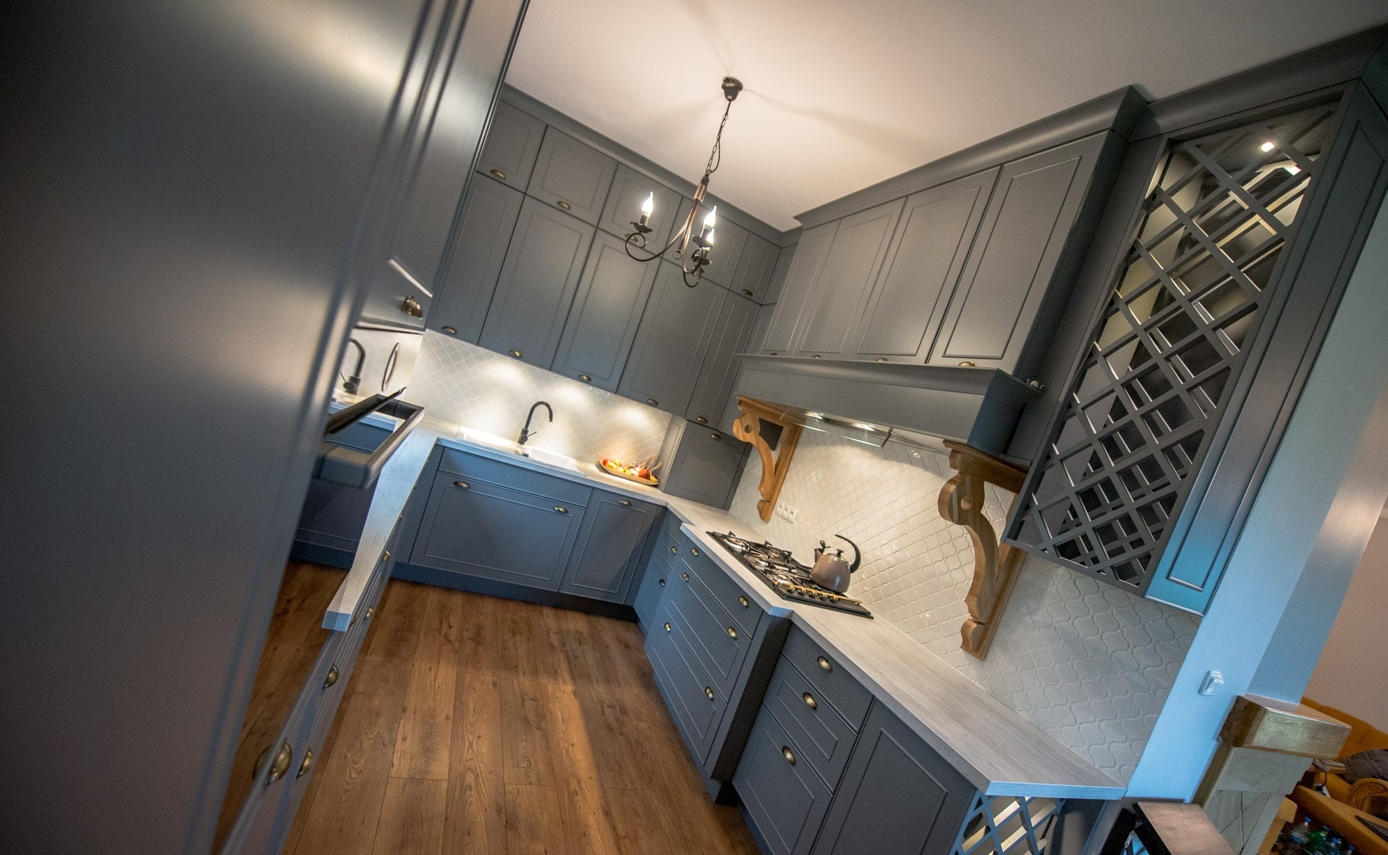 Szara, klasyczna kuchnia z drewnianą podłogą i dekoracyjną kratką nad blatem. Uwagę zwraca stylizowany okap z rzeźbionymi wspornikami i ozdobny żyrandol.