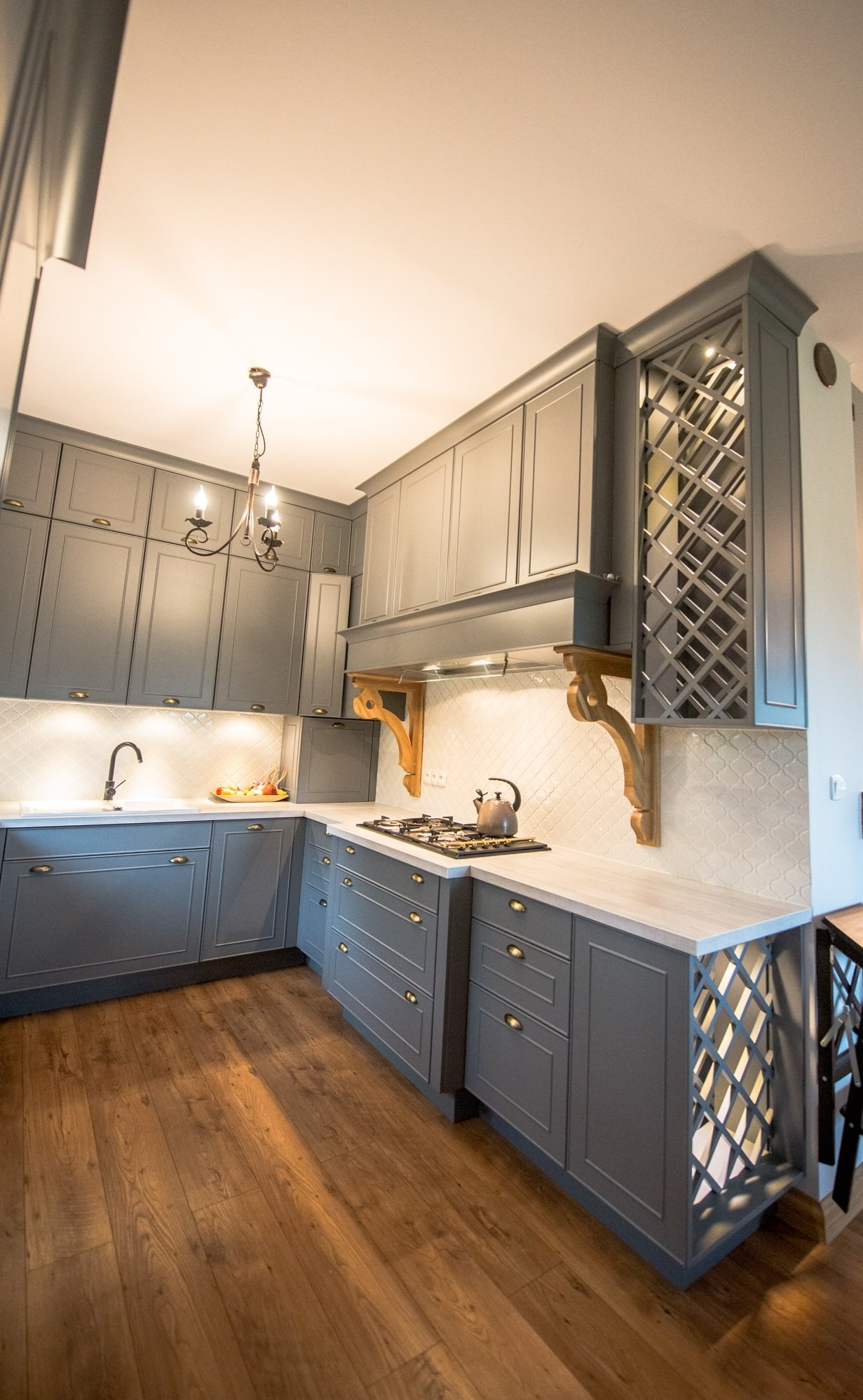 Szara kuchnia z frezowanymi frontami, drewnianym okapem i dekoracyjną kratką na szafce. Na blacie czajnik, oświetlenie punktowe, drewniana podłoga.