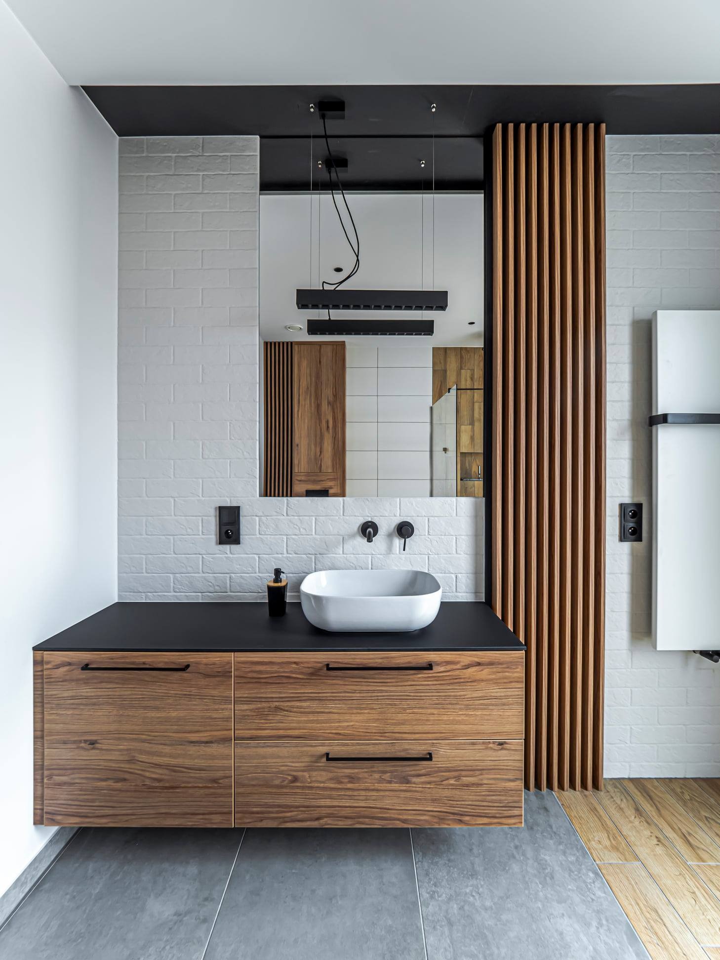 Nowoczesna łazienka z drewnianą szafką pod umywalką, czarnym blatem i dekoracyjnymi lamelami. Minimalistyczny design z białymi cegiełkami i lustrem.