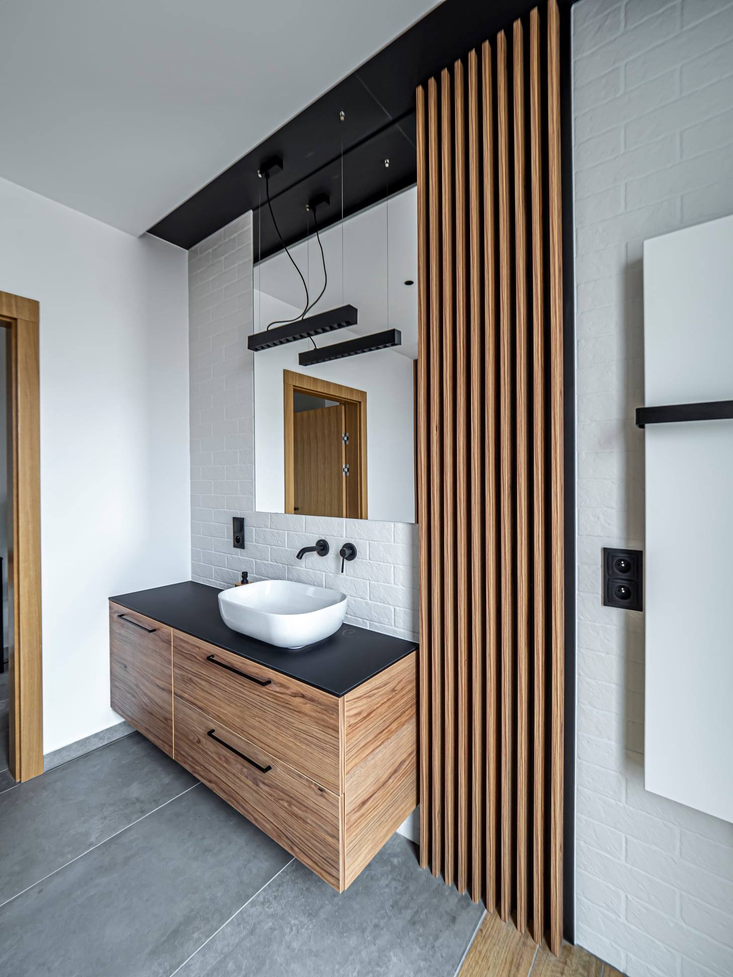 Nowoczesna łazienka z drewnianą szafką pod umywalką, białymi cegiełkami na ścianie i dekoracyjnymi drewnianymi lamelami. Minimalistyczne oświetlenie.