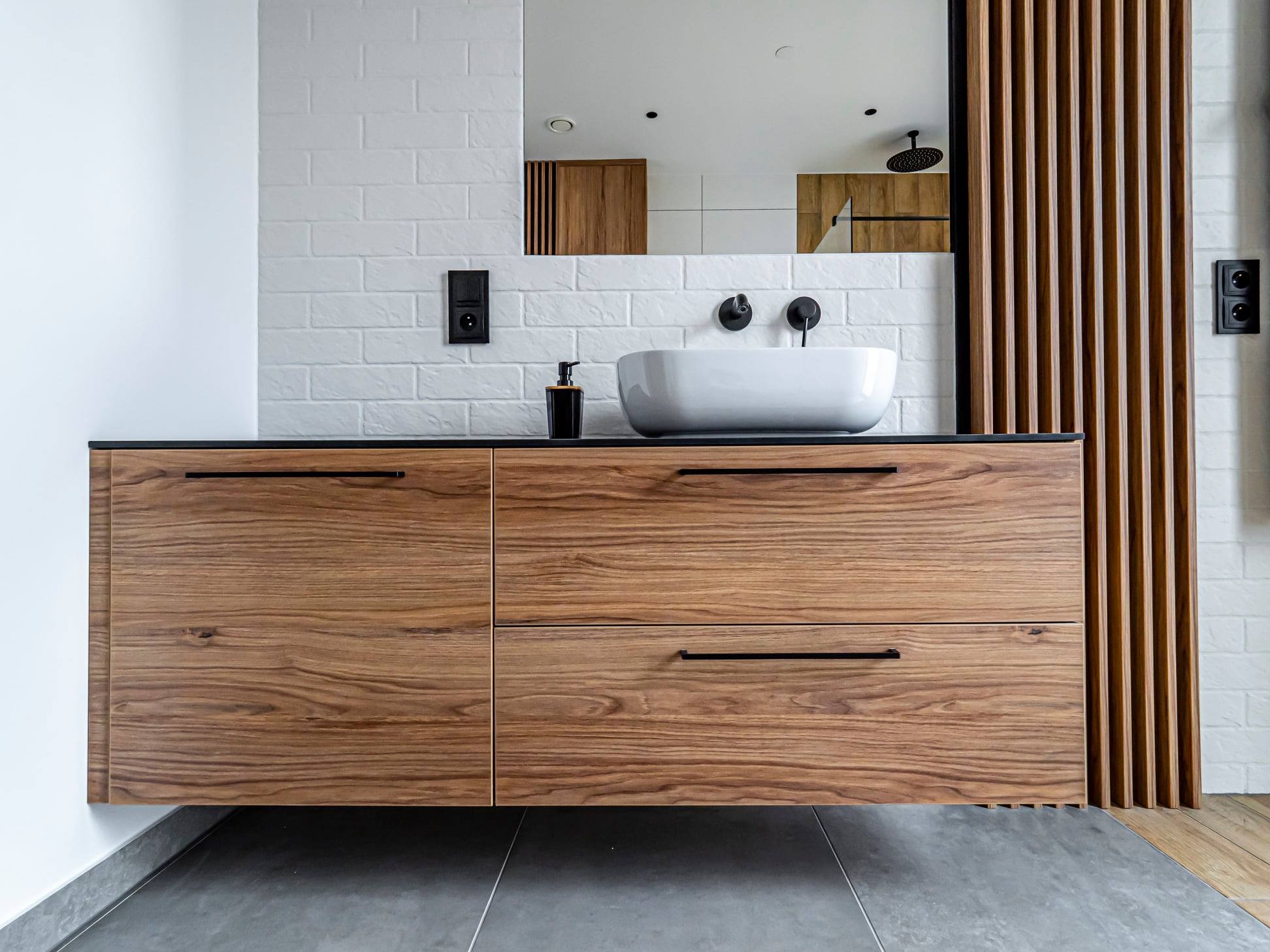 Drewniana szafka łazienkowa z umywalką nablatową i czarnymi detalami na tle białej cegły i drewnianej ścianki działowej. Minimalistyczny design.