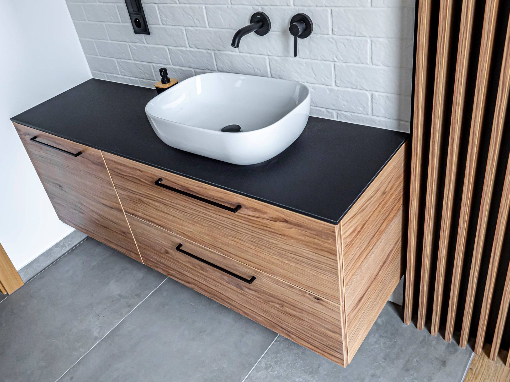 Nowoczesna szafka łazienkowa z drewna z czarnym blatem i białą umywalką nablatową. Minimalistyczna armatura w kolorze czarnym. Na ścianie biała cegła.